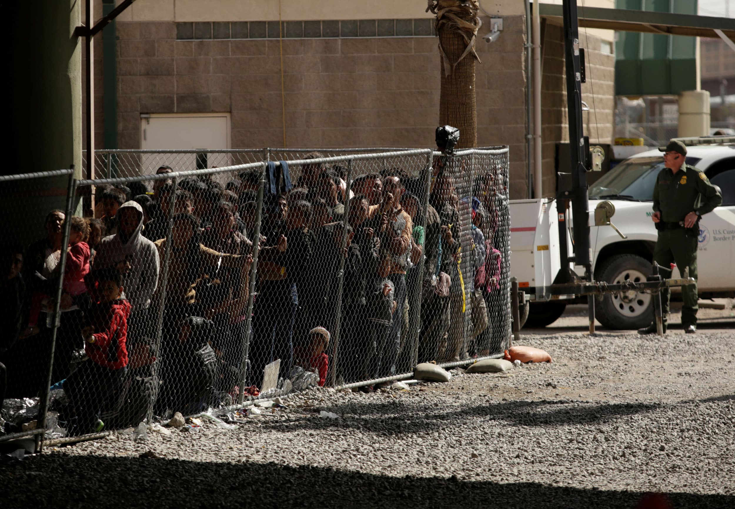 A group of migrants are shown enclosed behind a chain-link fence under an overpass.