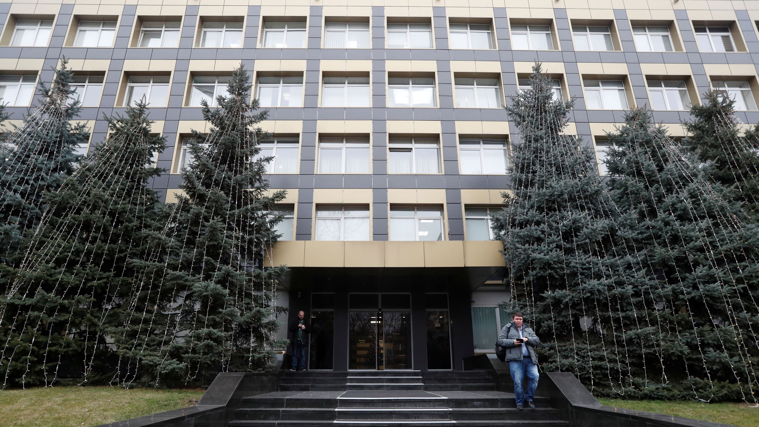 A large office building is shown with pine trees out front and a person walking down the stairs.