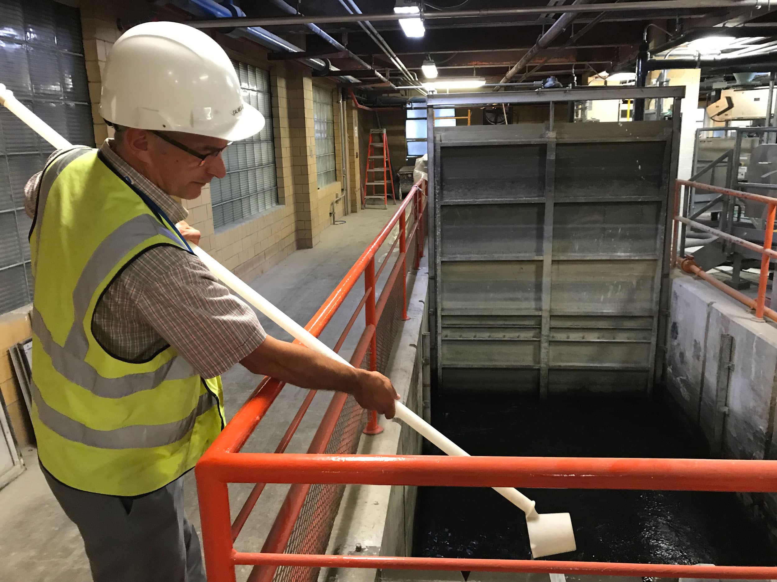 A man is shown wearing a safety vest and hard hat while holding a long white pole.