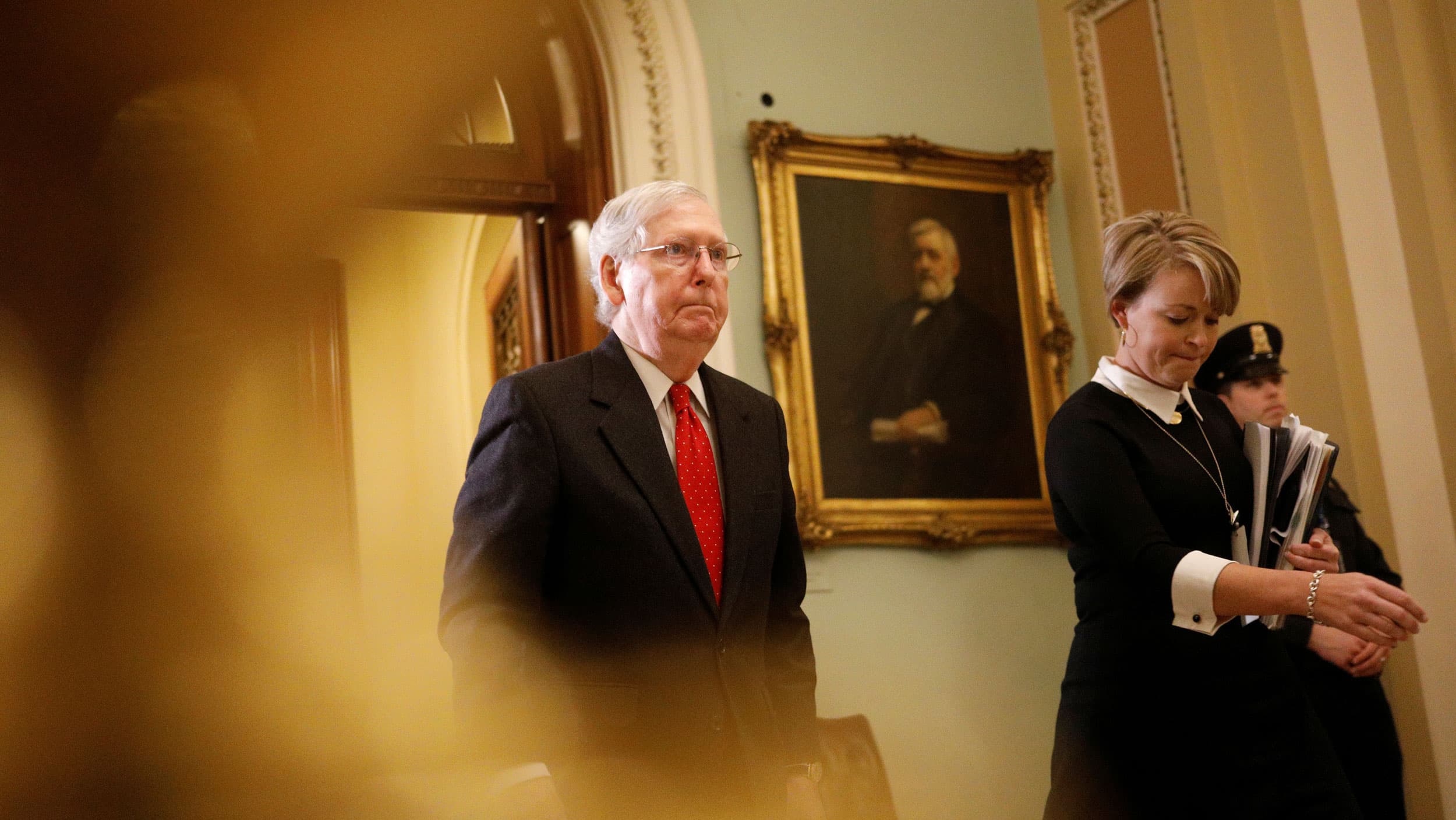 US Senate Majority Leader Mitch McConnell is shown wearing a dark suit and red tied walking.