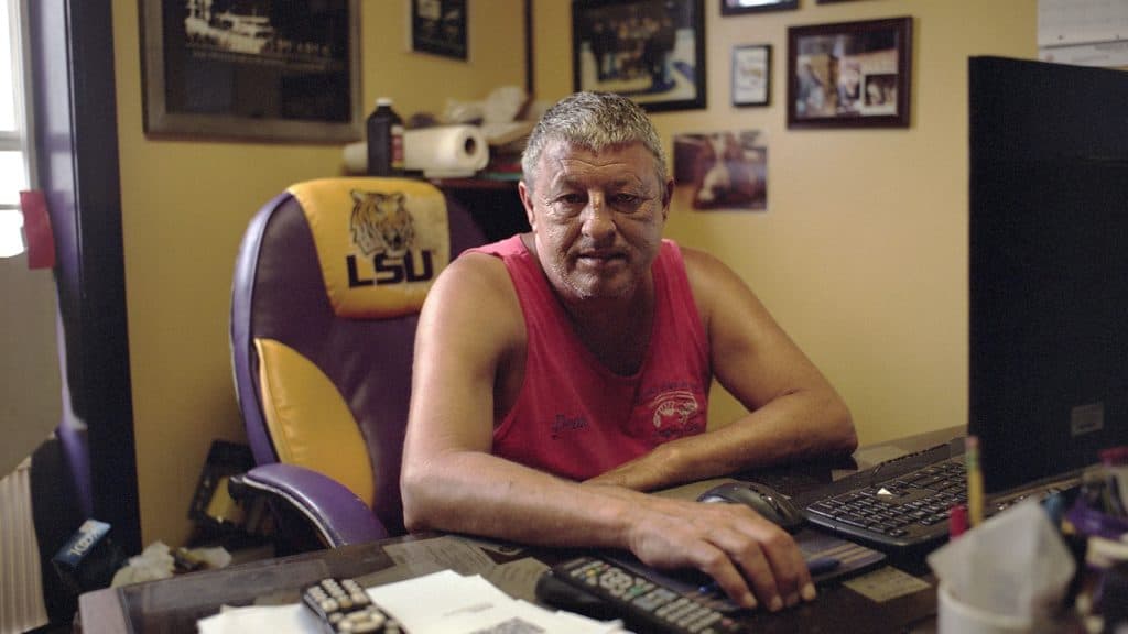 A man is shown sitting at a desk and wearing a red sleeveless shirt.