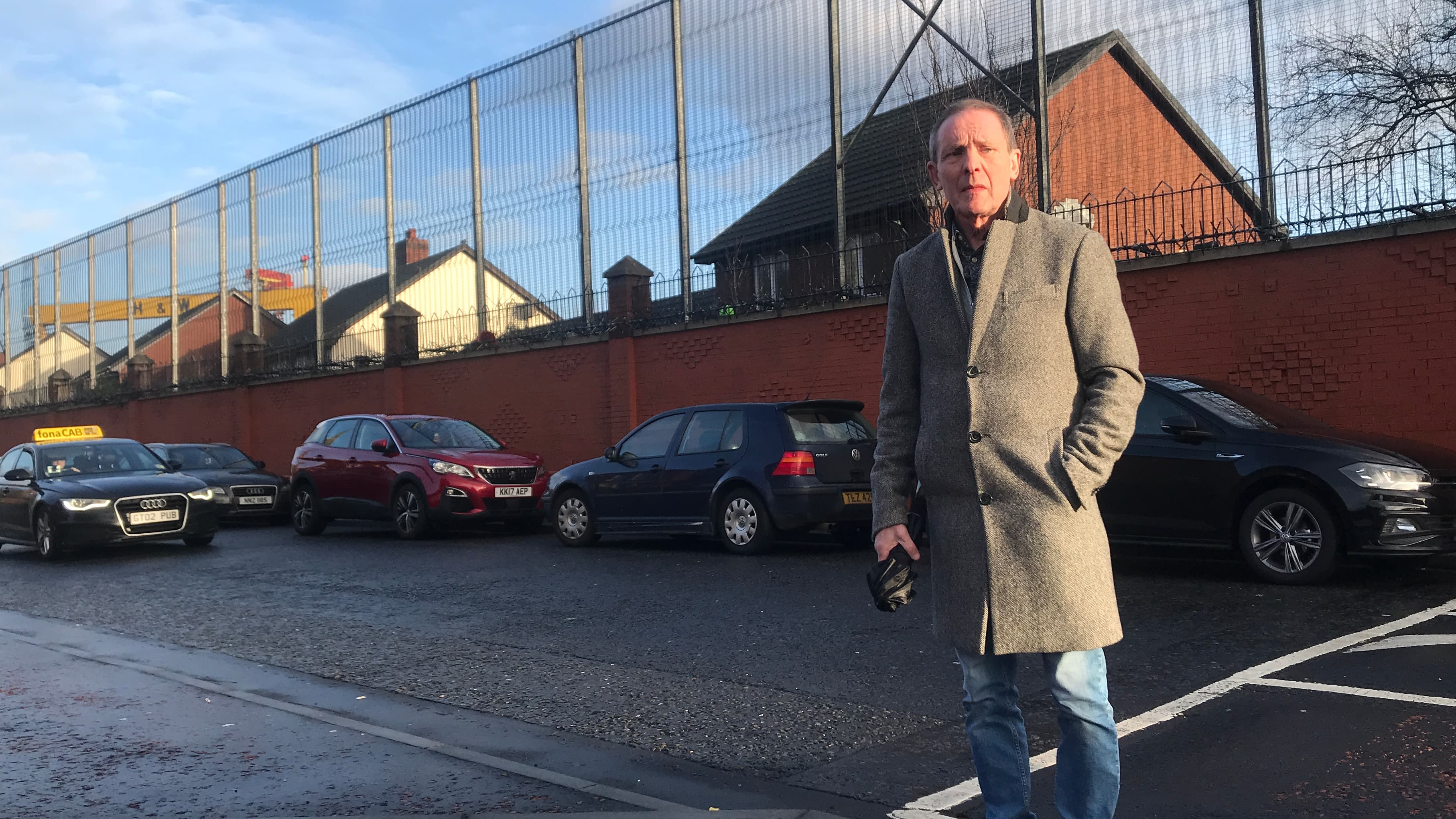 A man with silver hair stands outside with backdrop of border wall behind him.
