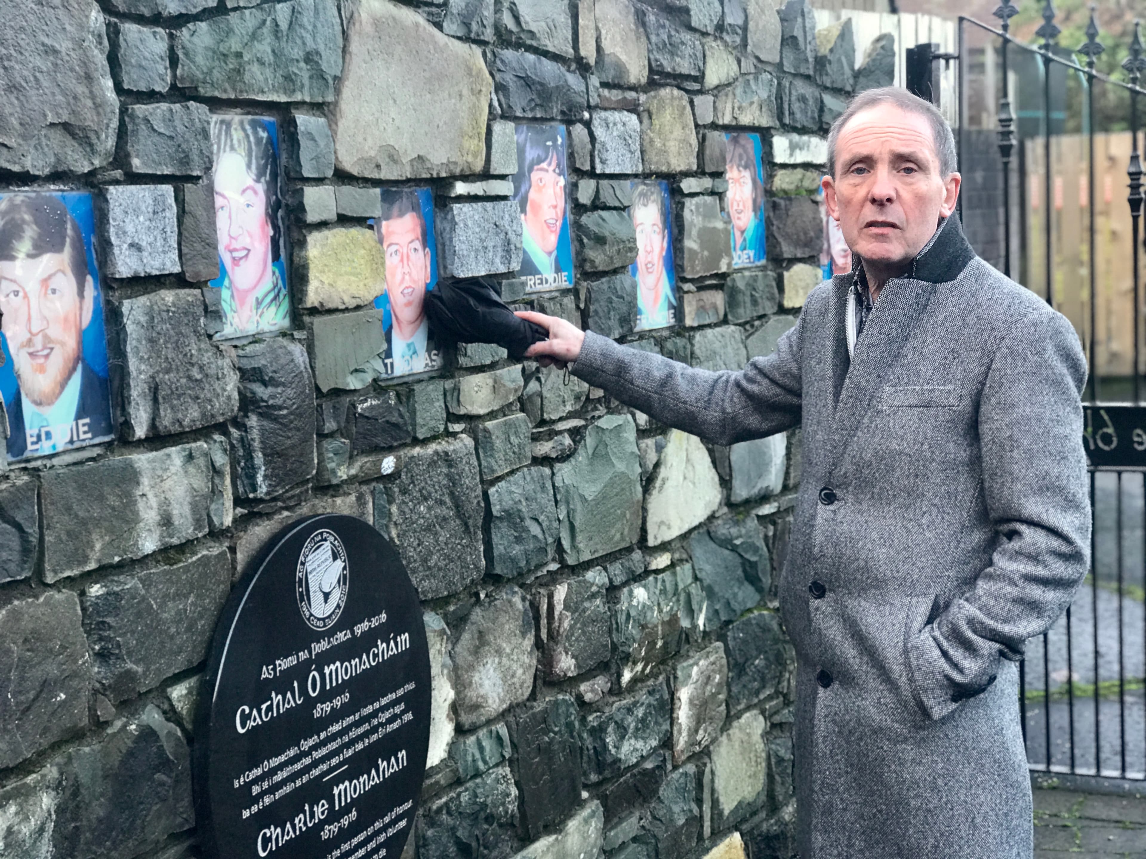 A man stands a wall with painted portraits and points to one.