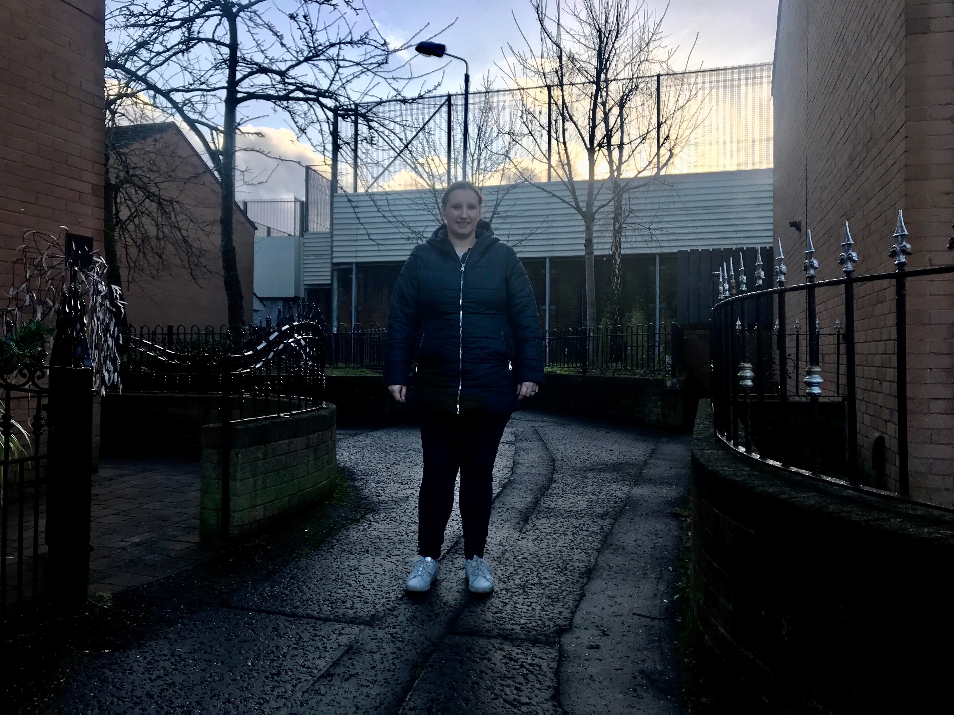 A woman stands with her hands in her pockets of her jacket with big border wall behind her.