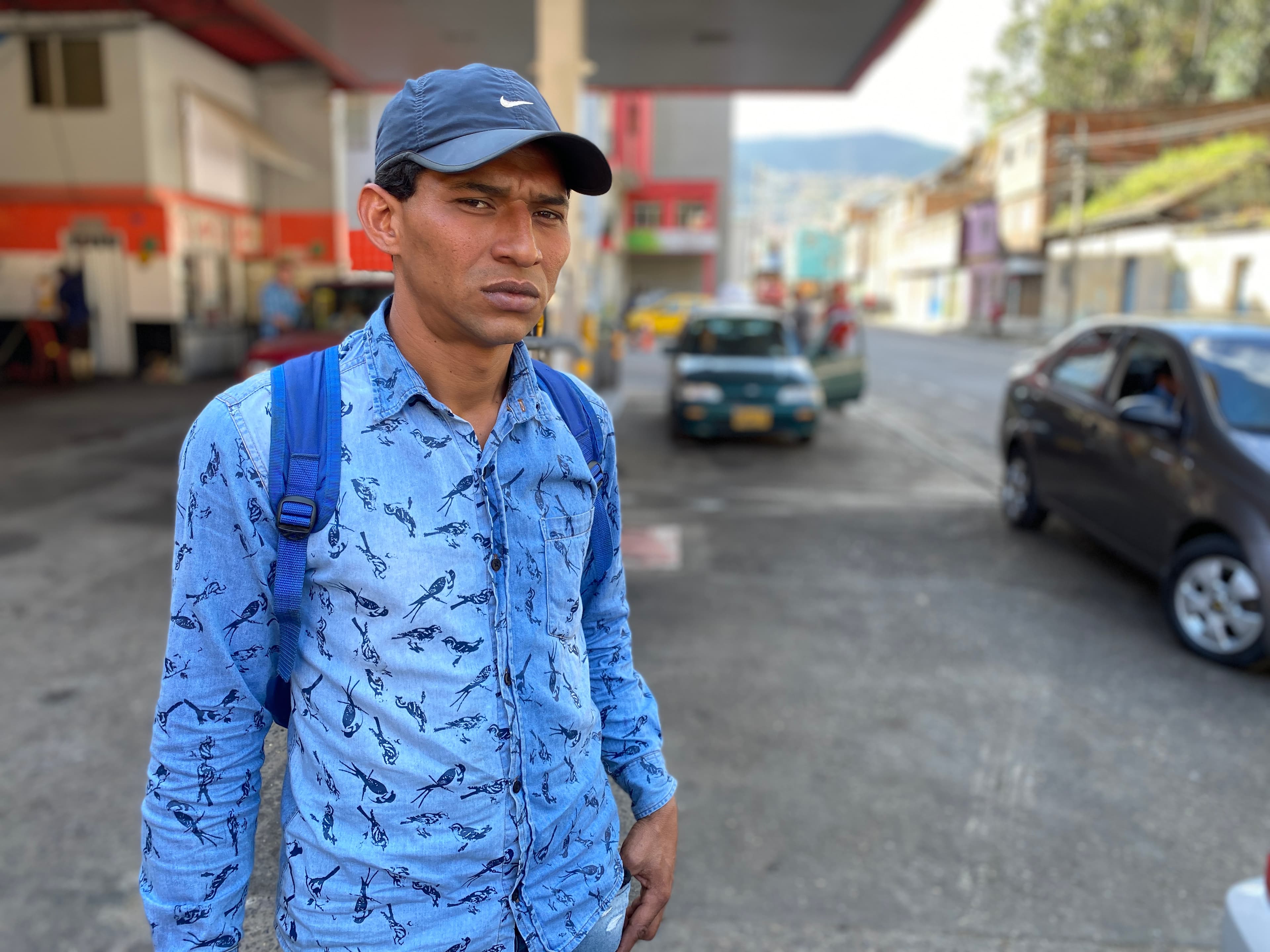 A man wears a light blue button down shirt, backpack and Nike cap at a gas station.