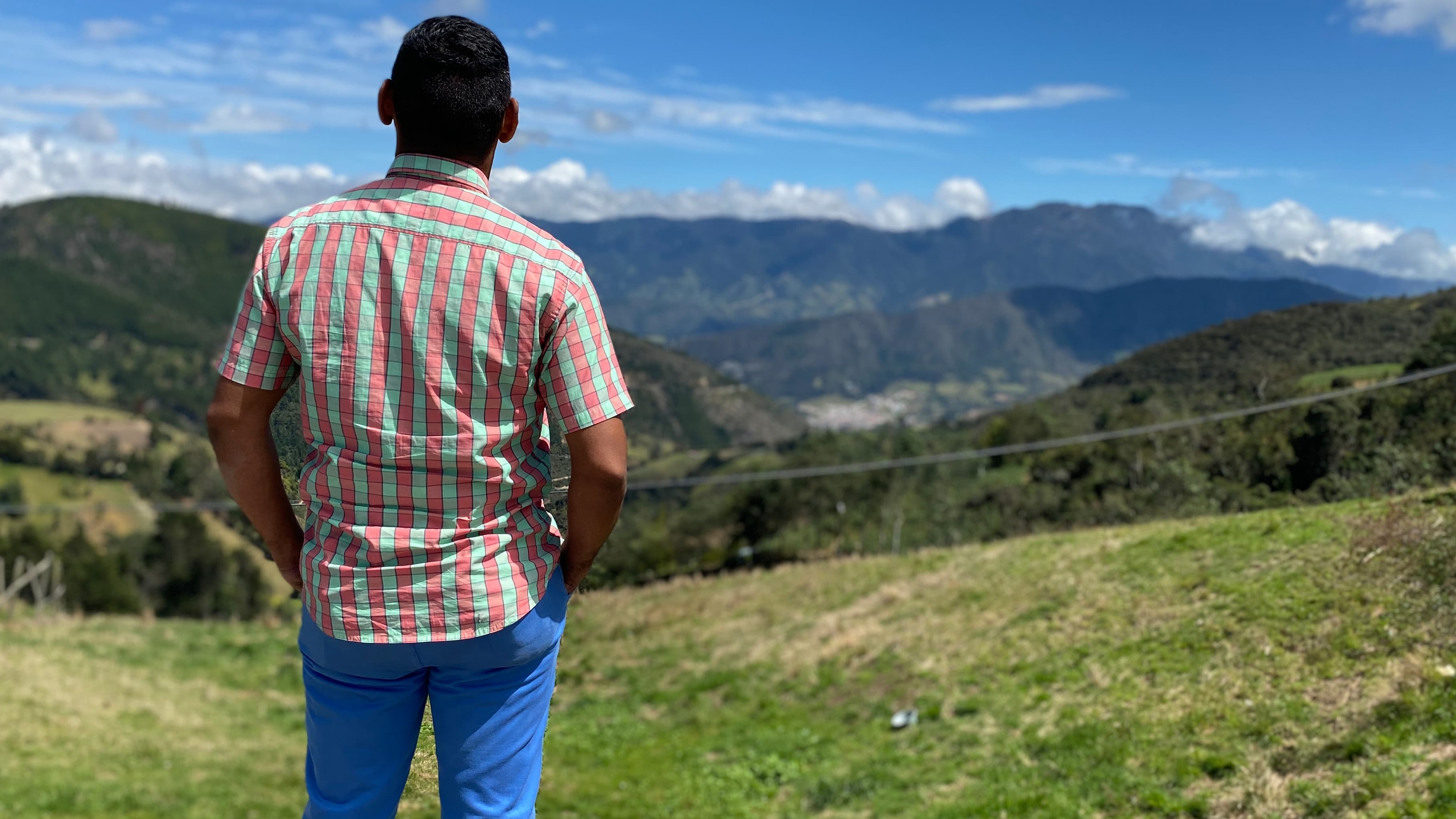 A man wears jeans and plaid shirt with back to camera facing mountain range