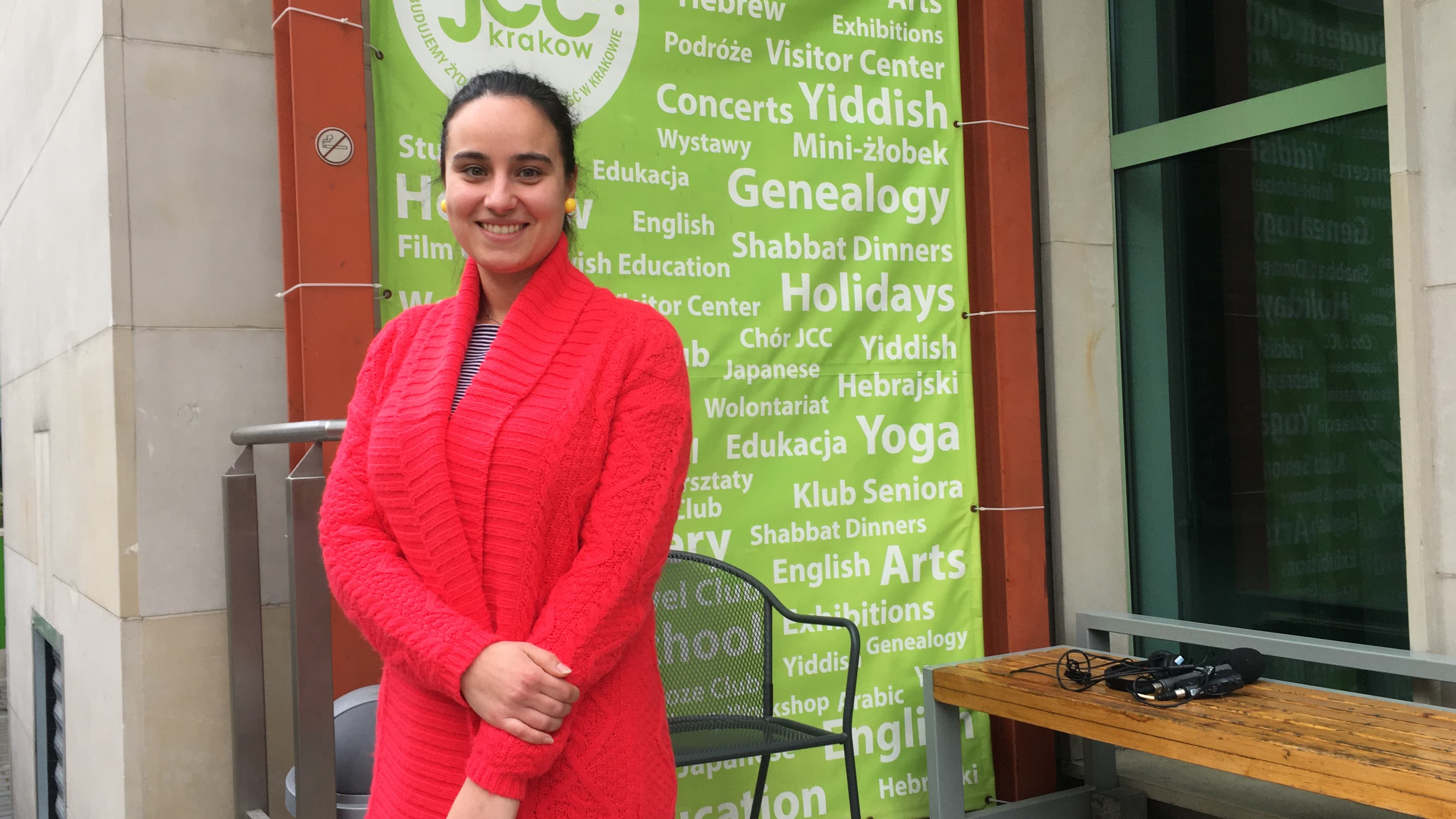 Marcjanna Kubala wears a red coat in front of a sign with Jewish-related words.