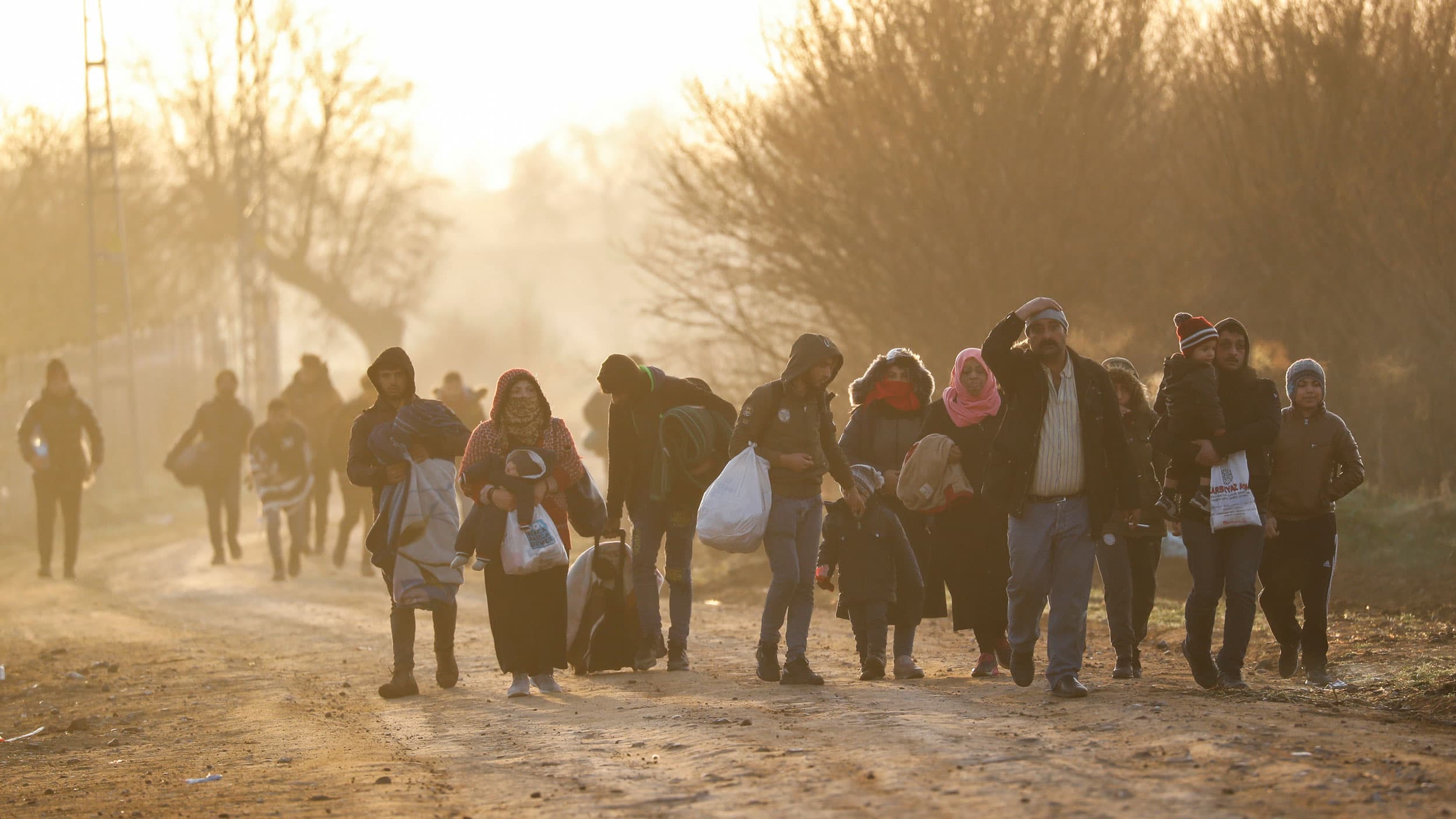A group of people are shown walking in a drit road and carrying their belonings.