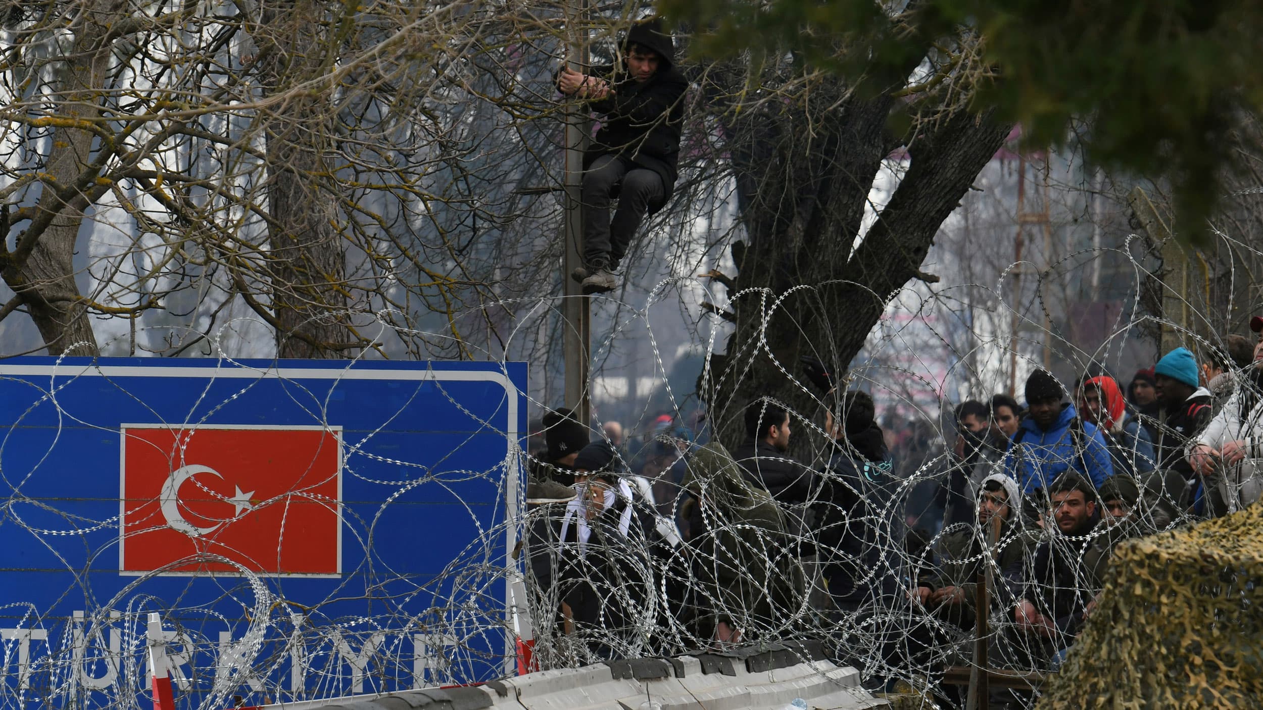 A group of migrants are shown standing together behind a border fence with razor wire.