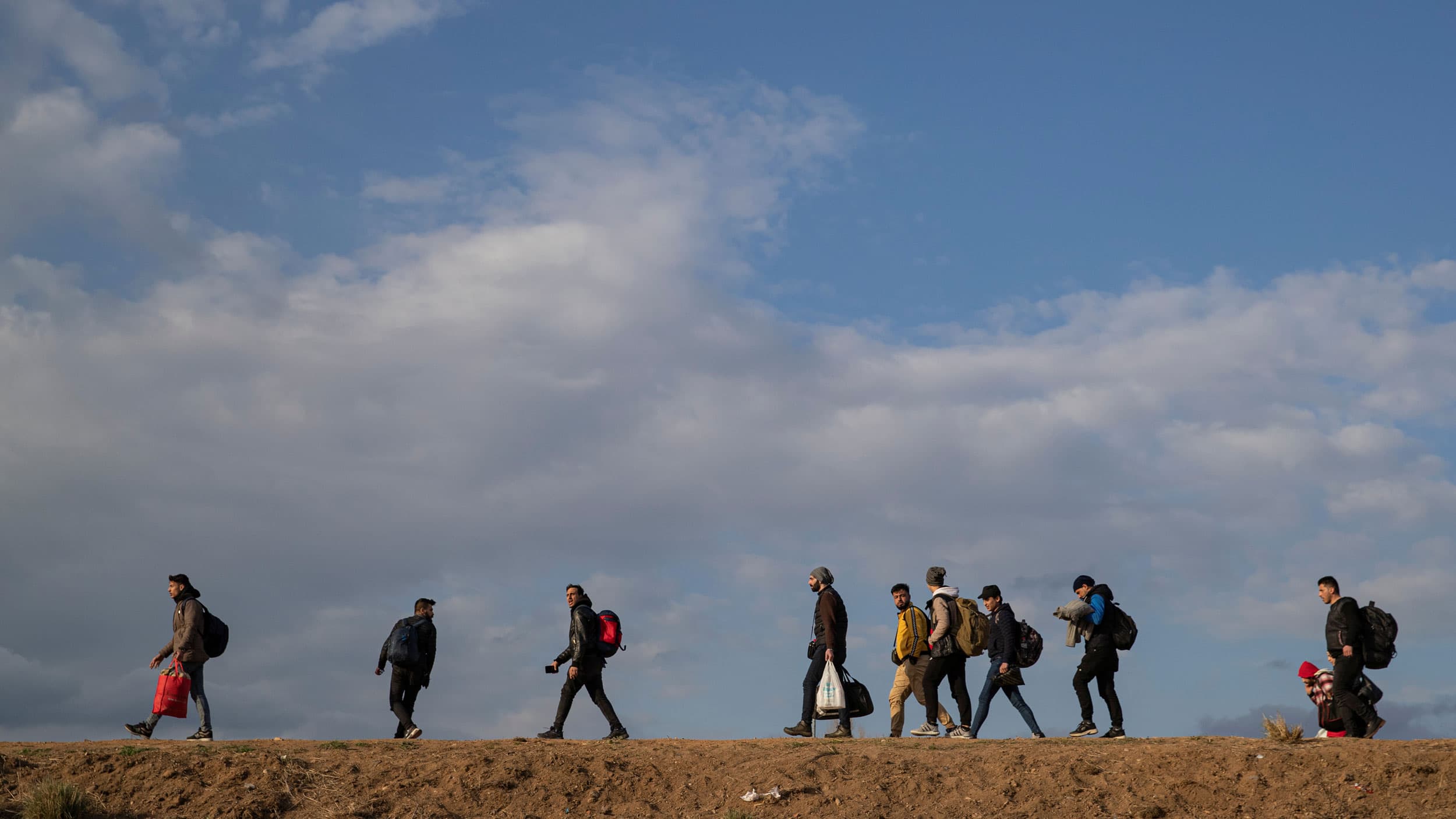 Several people are shown from a distance walking in a line and carrying their belongings.