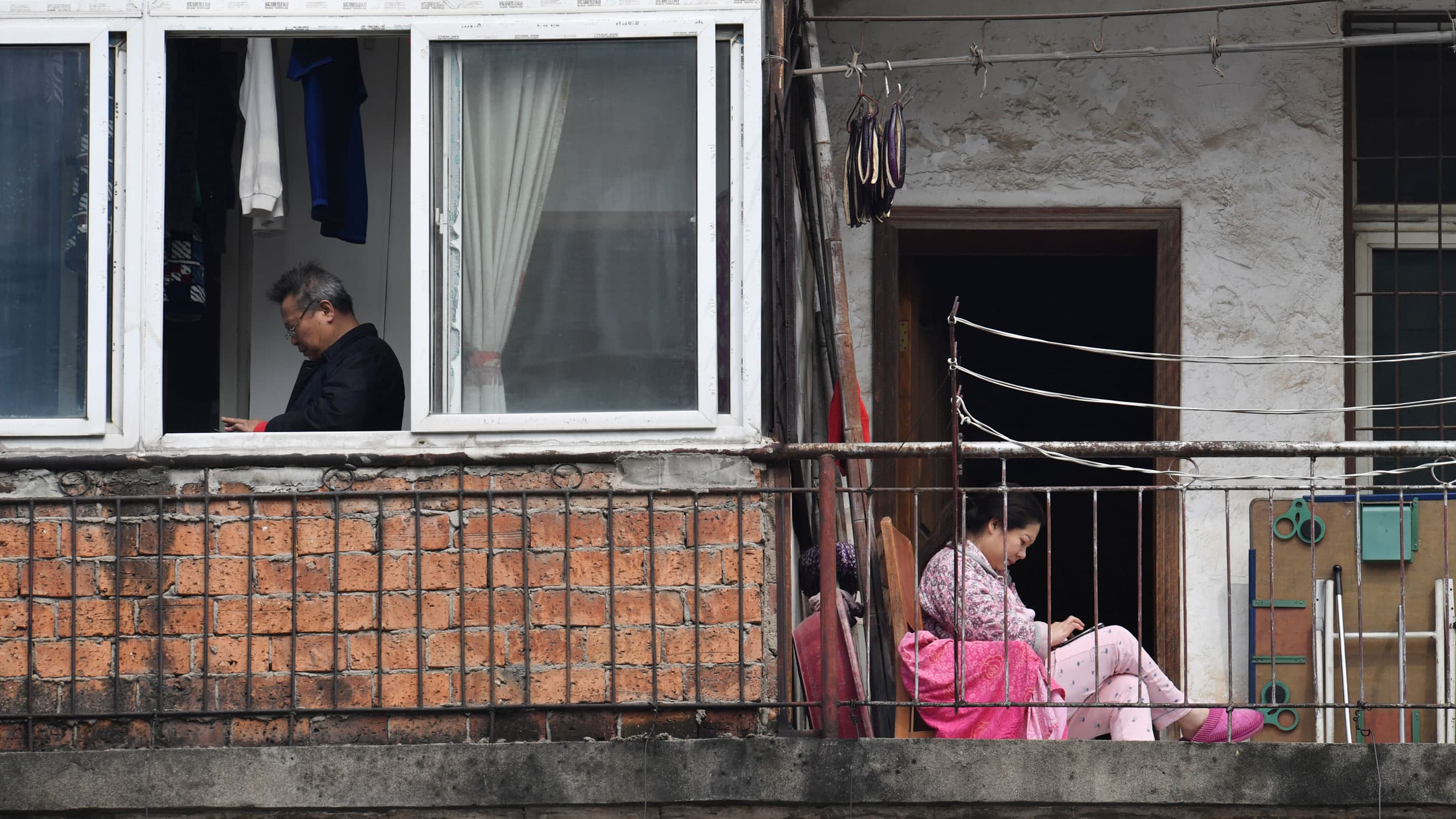 A woman is shown sitting and wearing pink while looking at her mobile phone adjacent to a man shown through an open window.