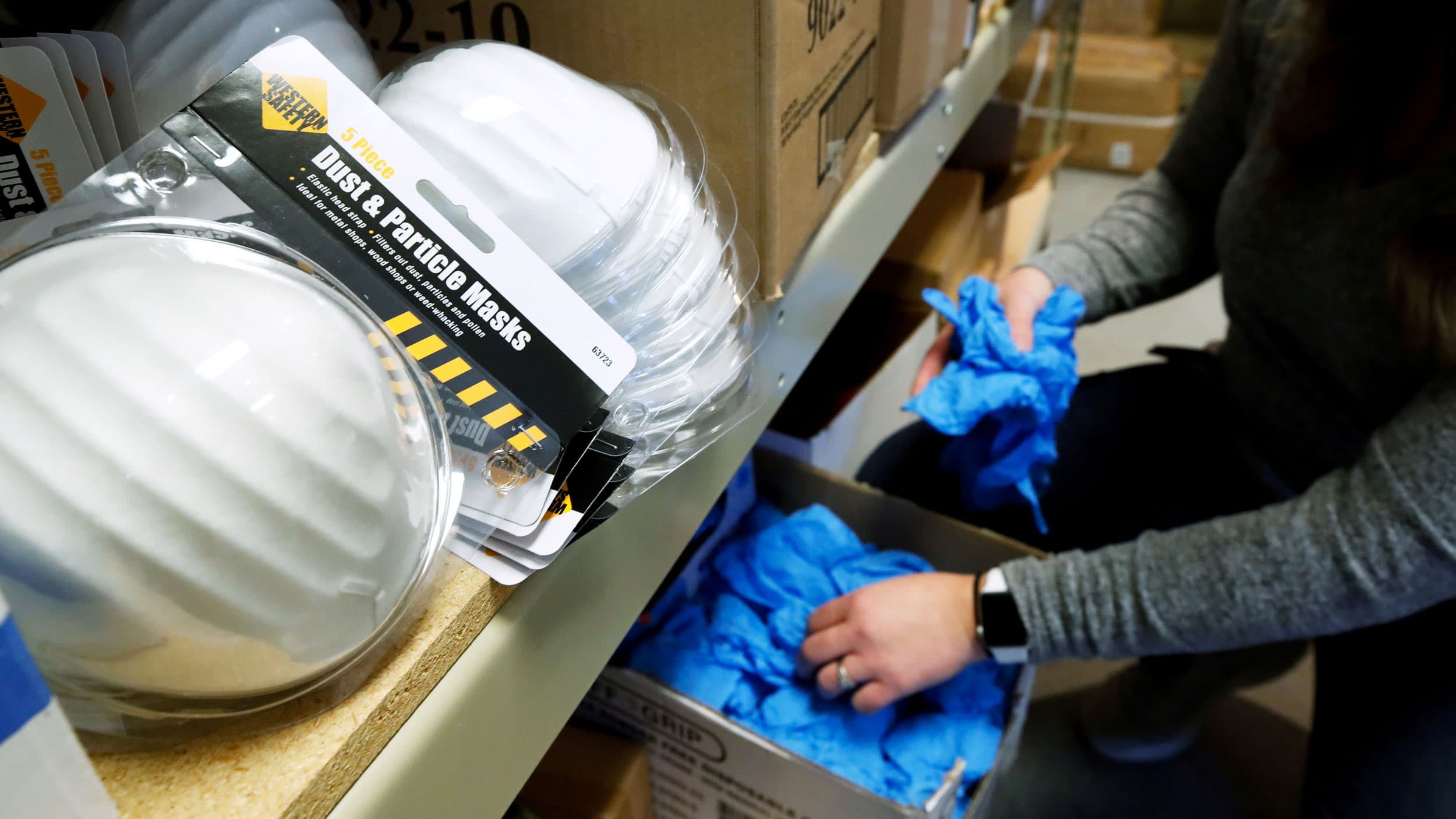 A woman grabs blue disposable gloves from a box as face masks sit in the frame.