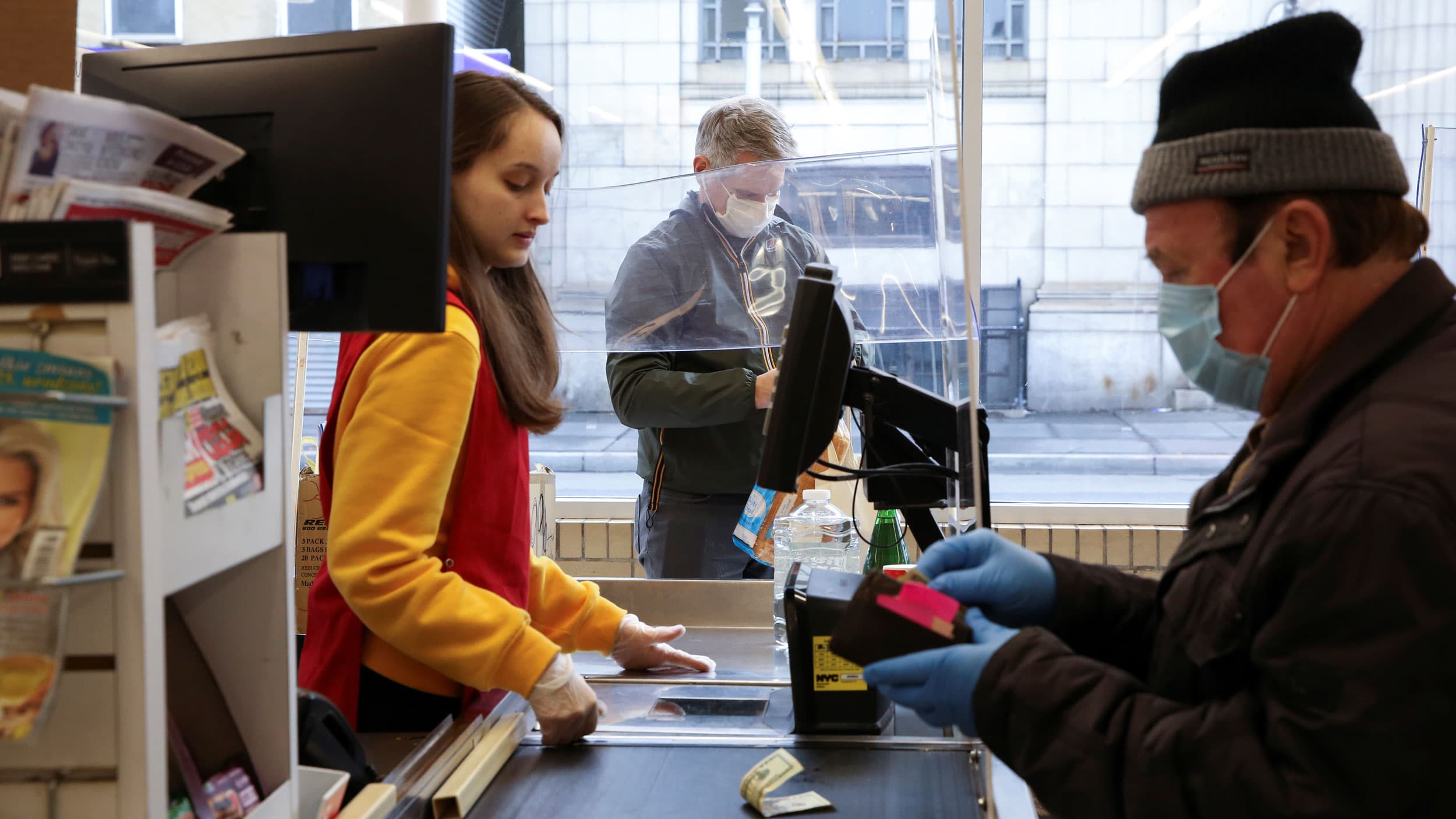 A man packs groceries separated from the cashier by a plastic sheet