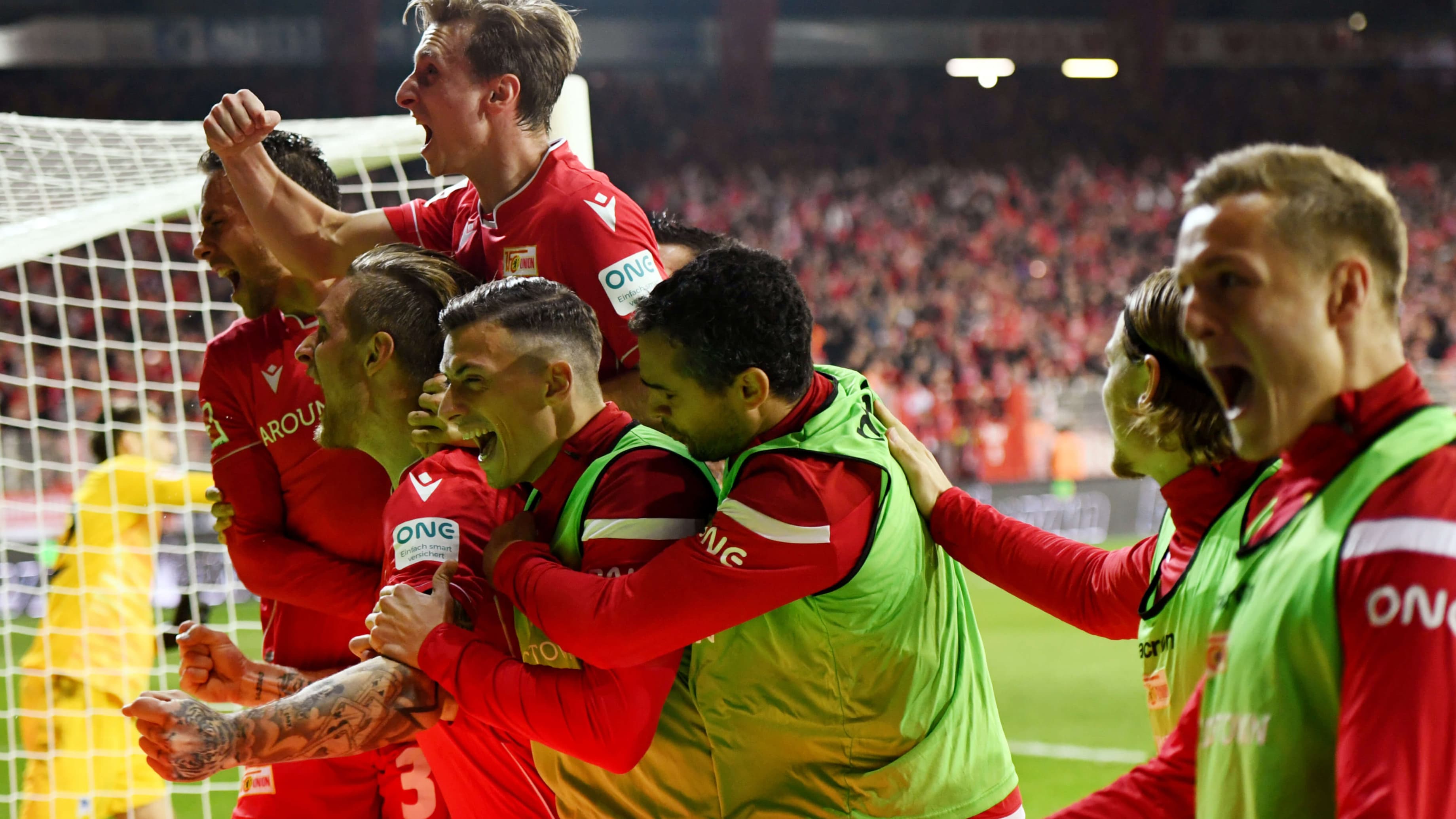A group of men in red uniforms jump for joy on a soccer field.
