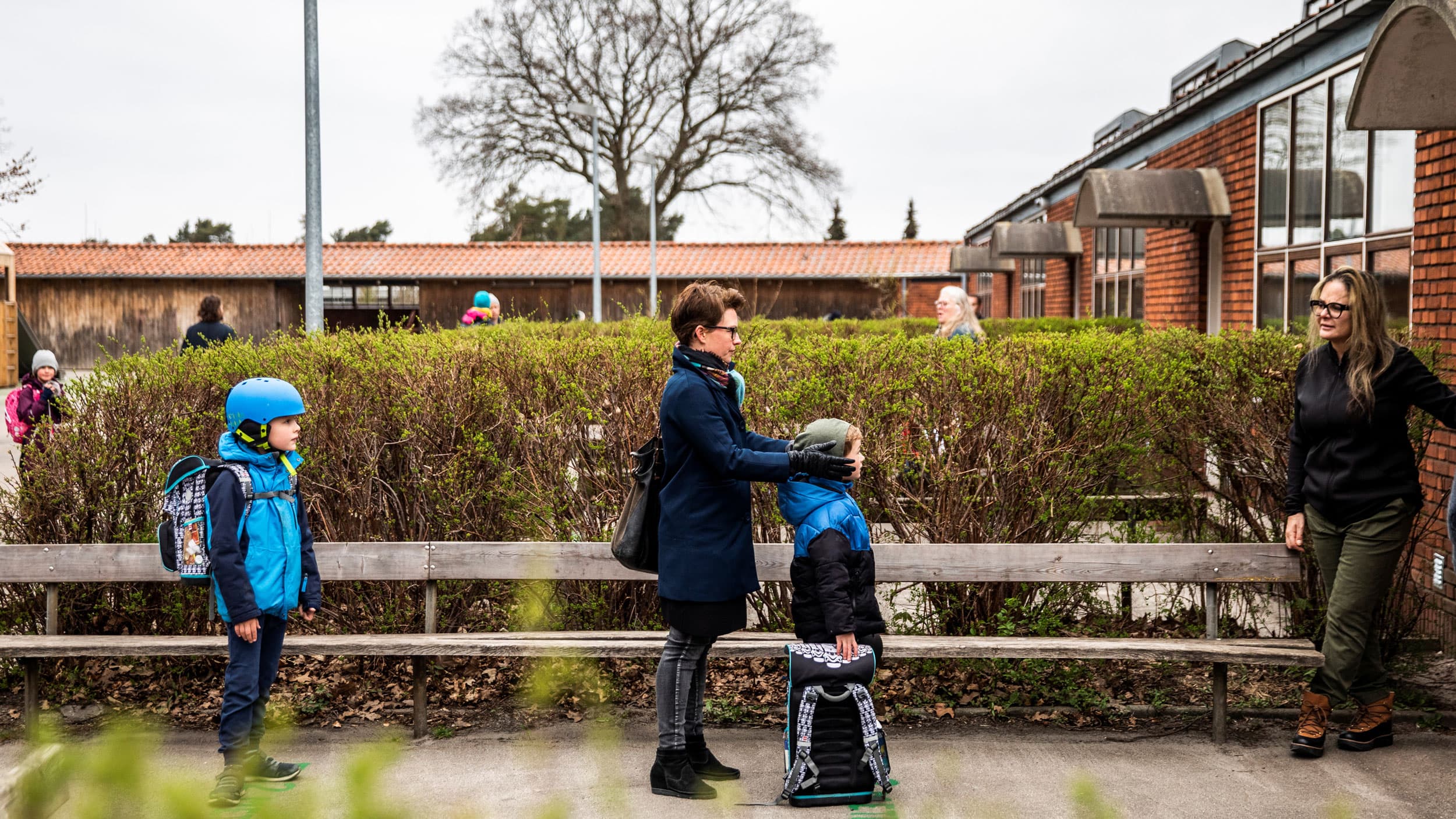 Several children and their parents are shown standing in a line spaced away from each other with a long wooden bench along side.