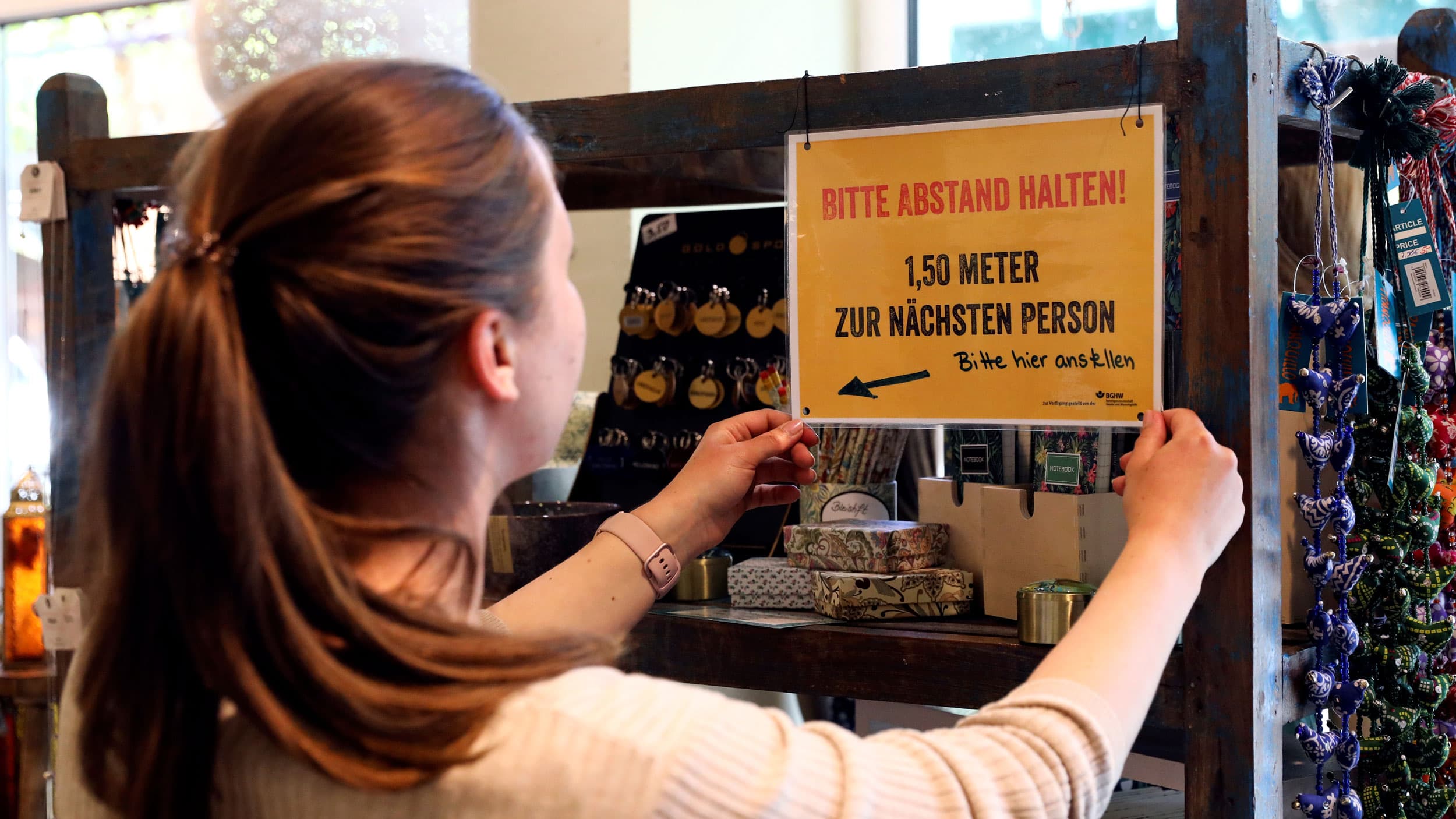 A woman with long hair is shown hanging a warning sign with an arrow on it cautioning patrons to keep their social distance.