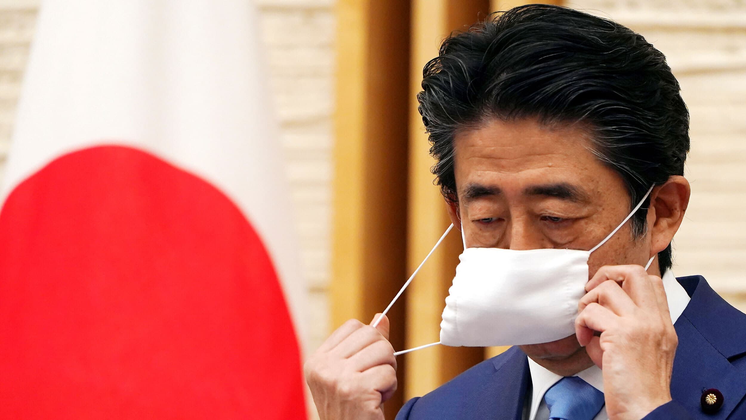 Japan's Prime Minister Shinzo Abe is shown removing a white protective face mask and wearing a blue suit with the Japanese flag in the distance.