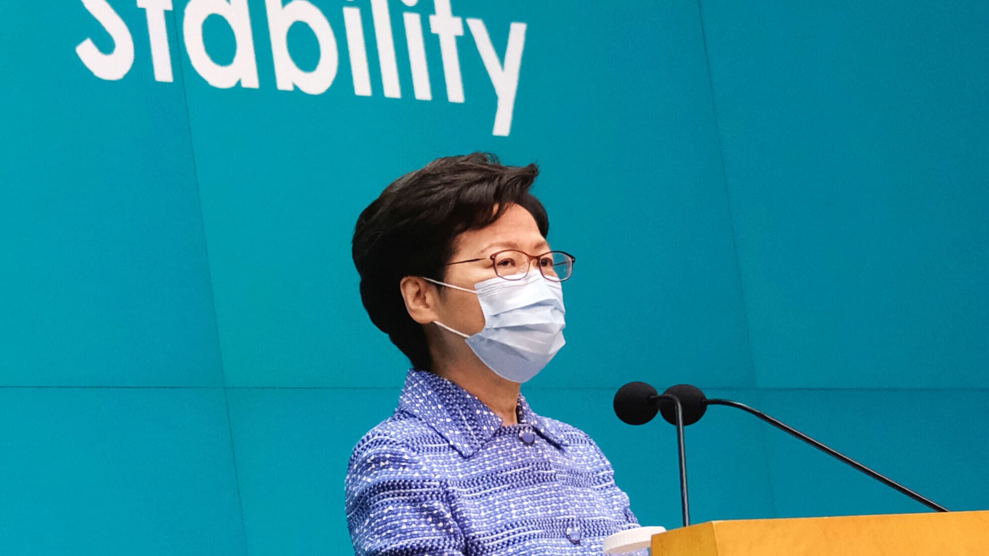 Hong Kong Chief Executive Carrie Lam is shown standing at a wooden podium with two small microphones and wearing a protective face mask.