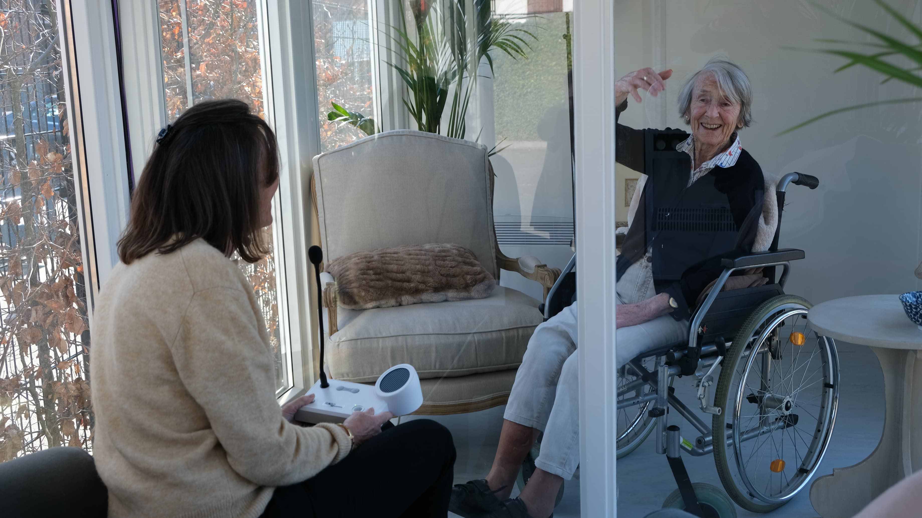 A younger woman looks at an older woman behind glass at The Glass Garden House at the Claris nursing home.