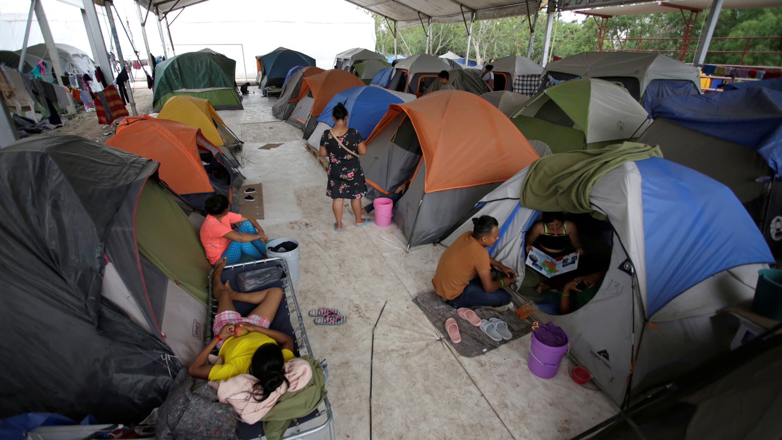 Several rows of dome tents are show under a large canopy with asylum seekers shown sitting outside their tents.