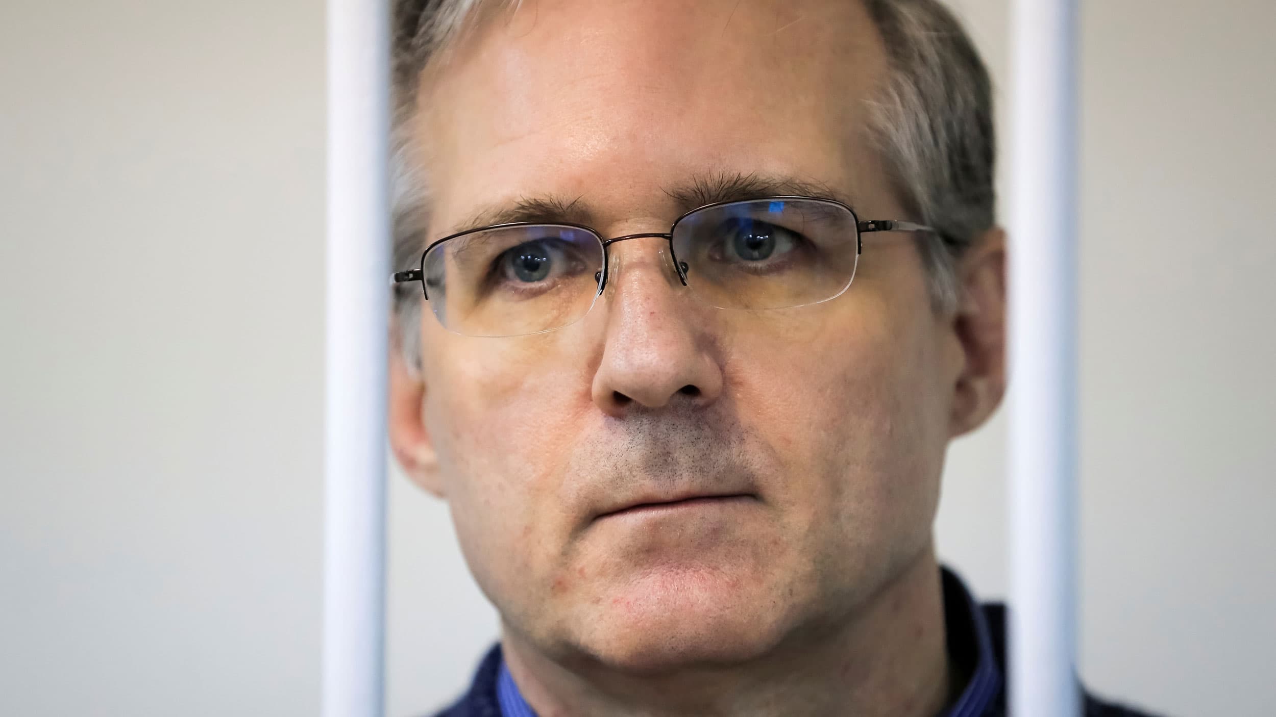 A close up photograph of Paul Whelan who is wearing glasses and is behind white metal bars.