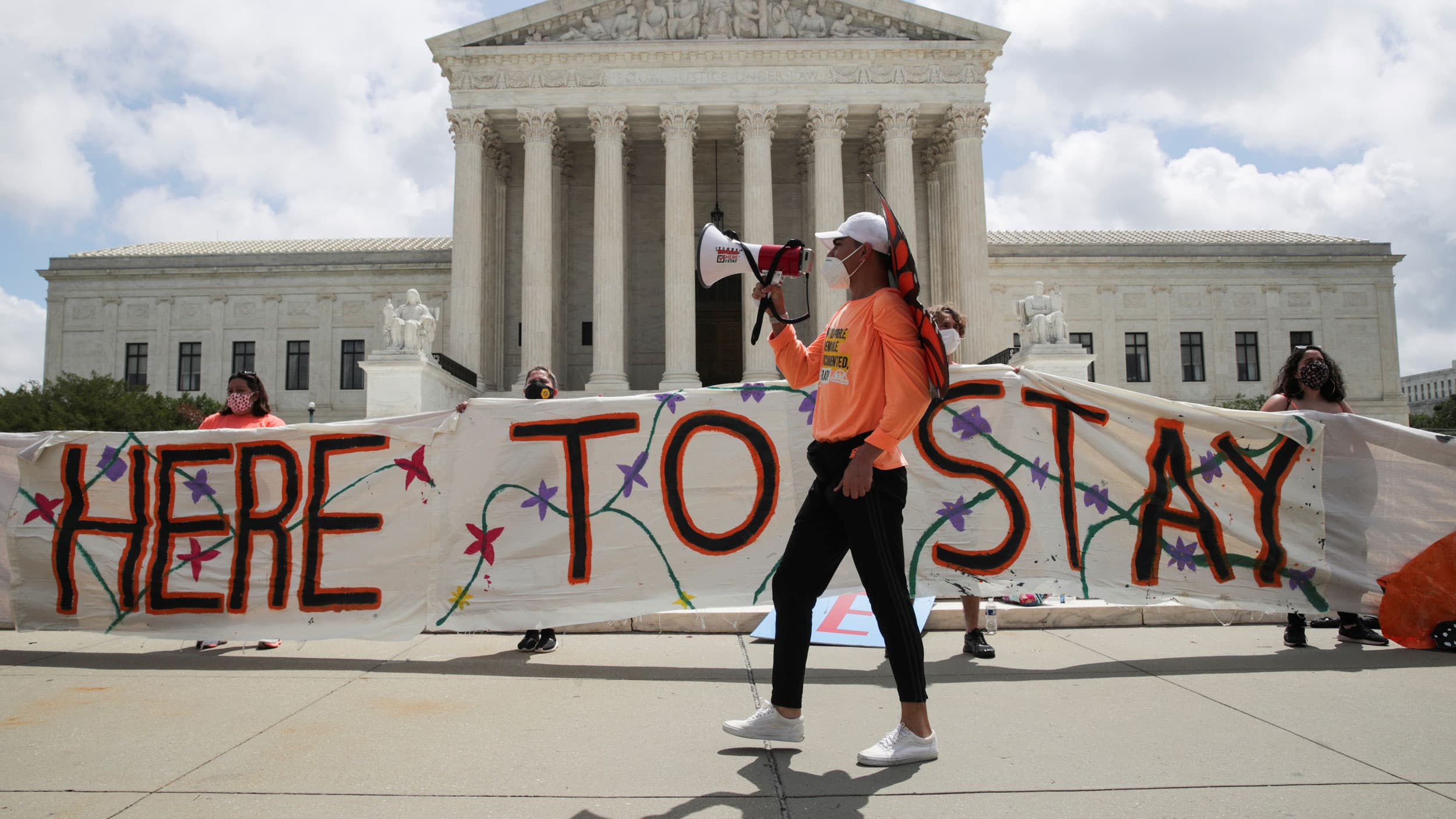 Several people are shown holding a large banner that says, 'Here To Stay