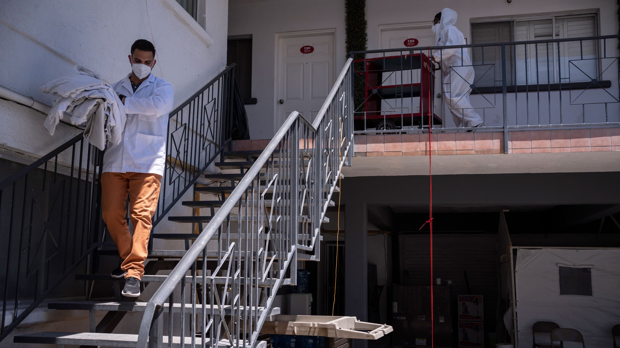 Two men are shown wearing protective medical gear and carrying various supplies while walking outside of Holtel Filtro.