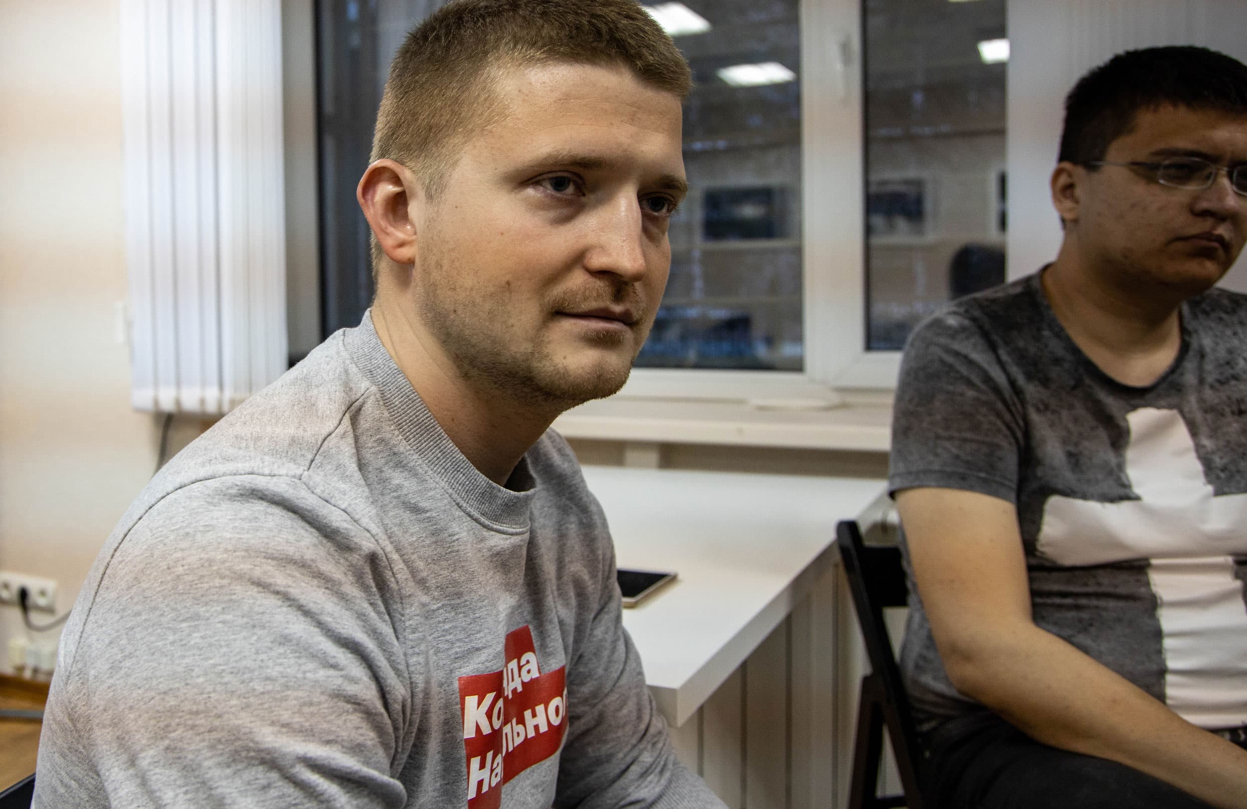 A young man sits next to another man in an office setting.