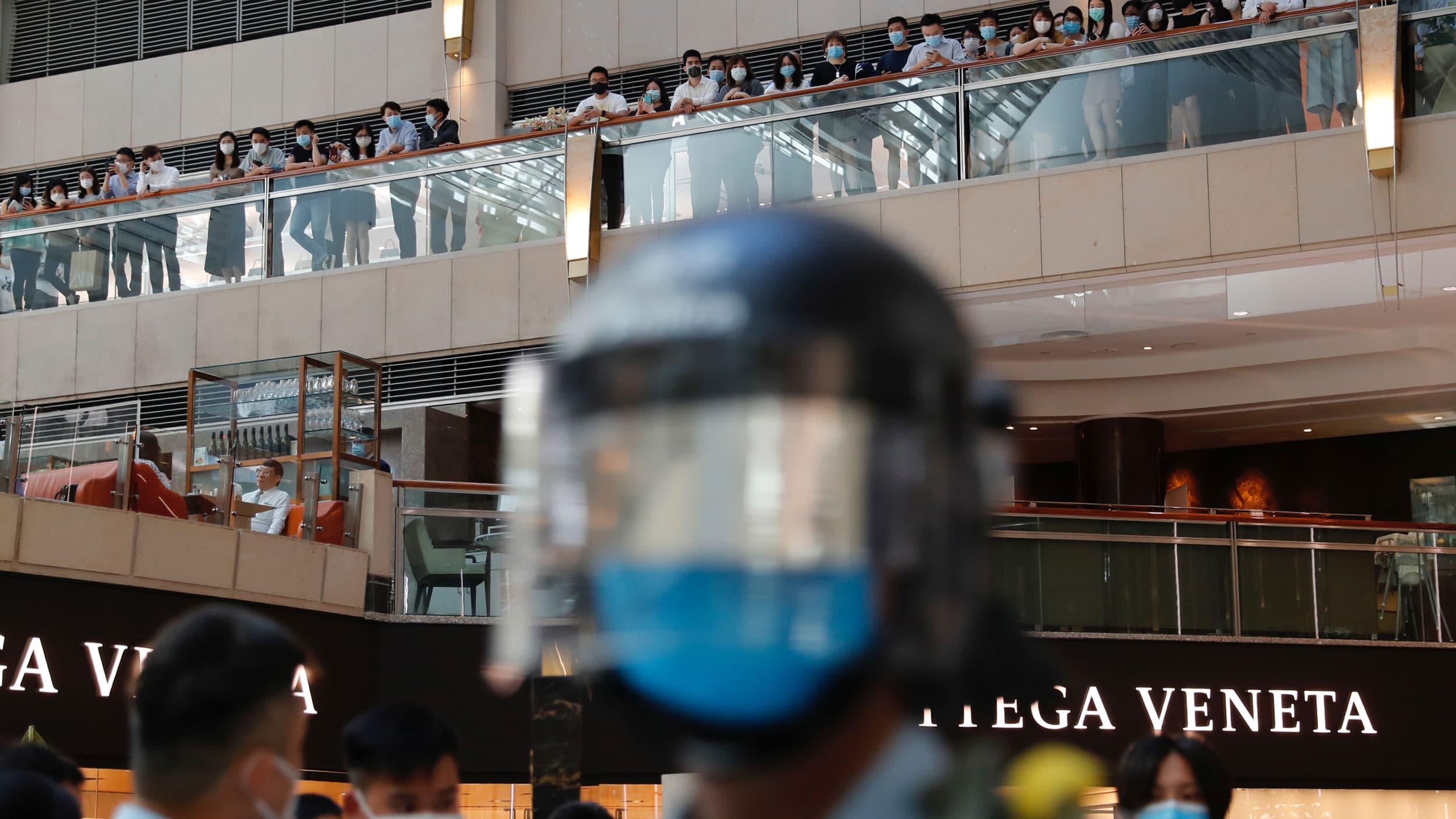 A helmeted head is blurry in the foreground, behind it, a line of protesters on a balcony
