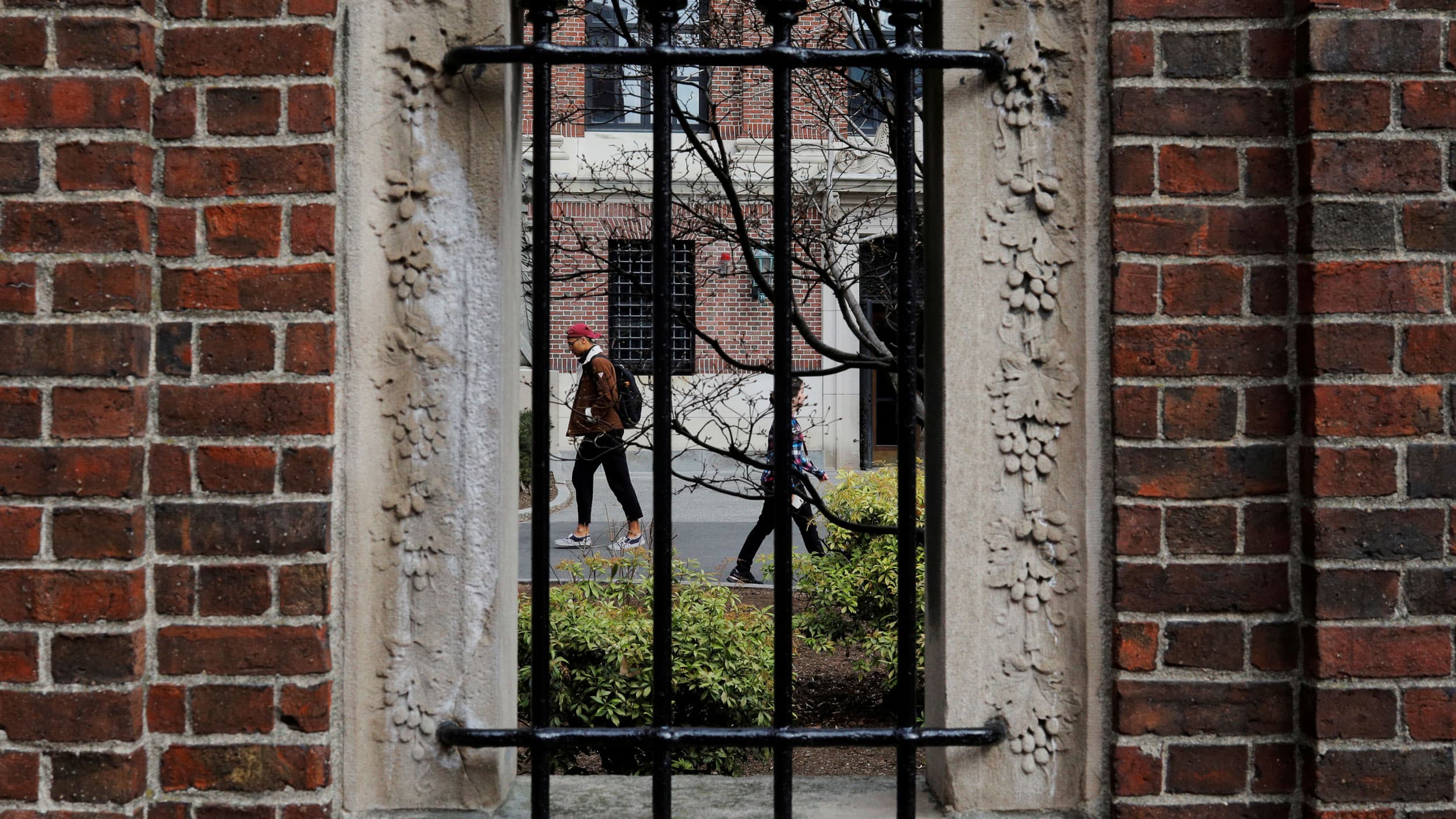 Several people are shown through a stone window with metal bars on it walking.
