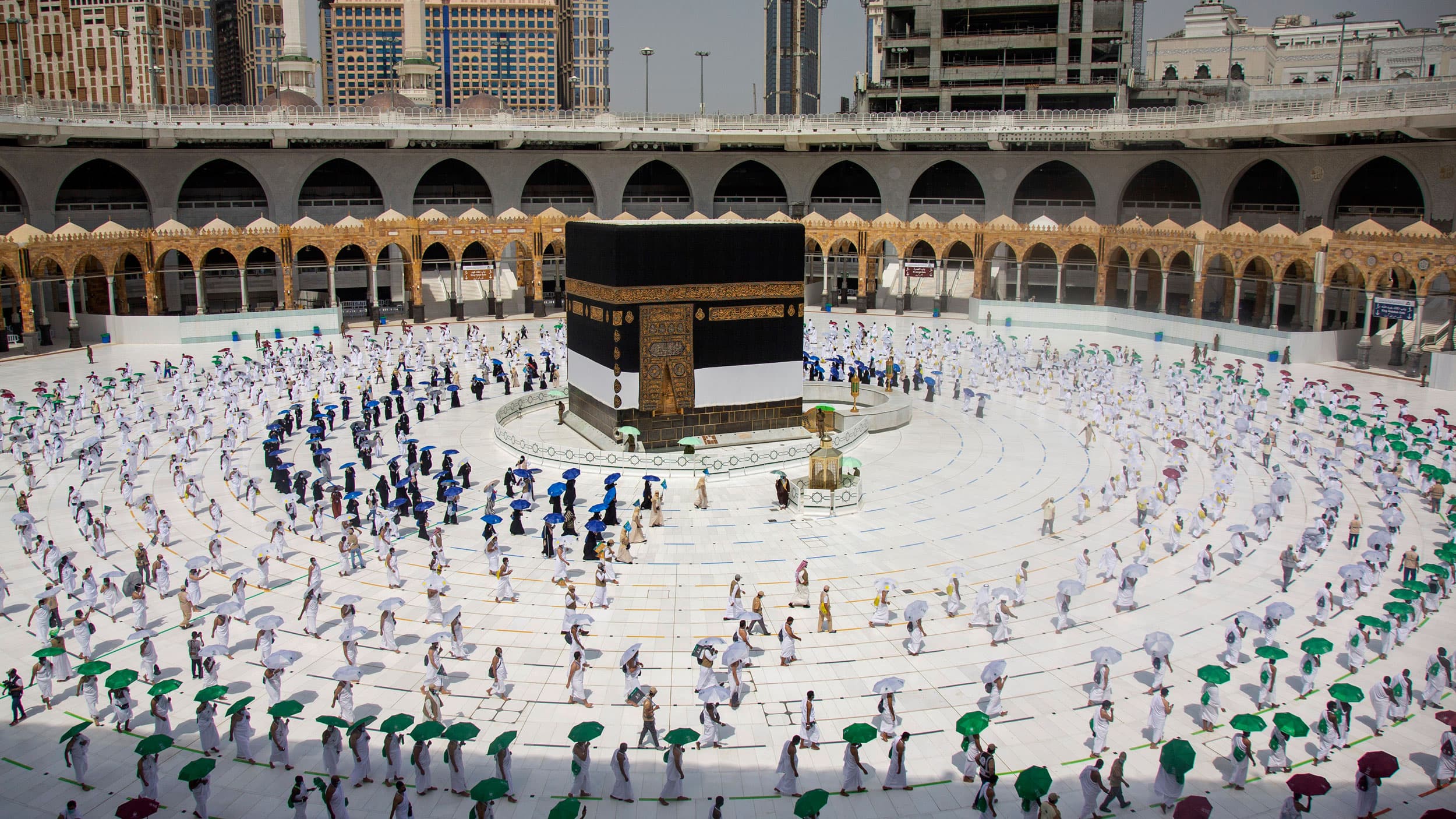 Pilgrims are shown from above around the cube-like Kaaba in Mecca walking on specified lines and carrying colored umbrellas.