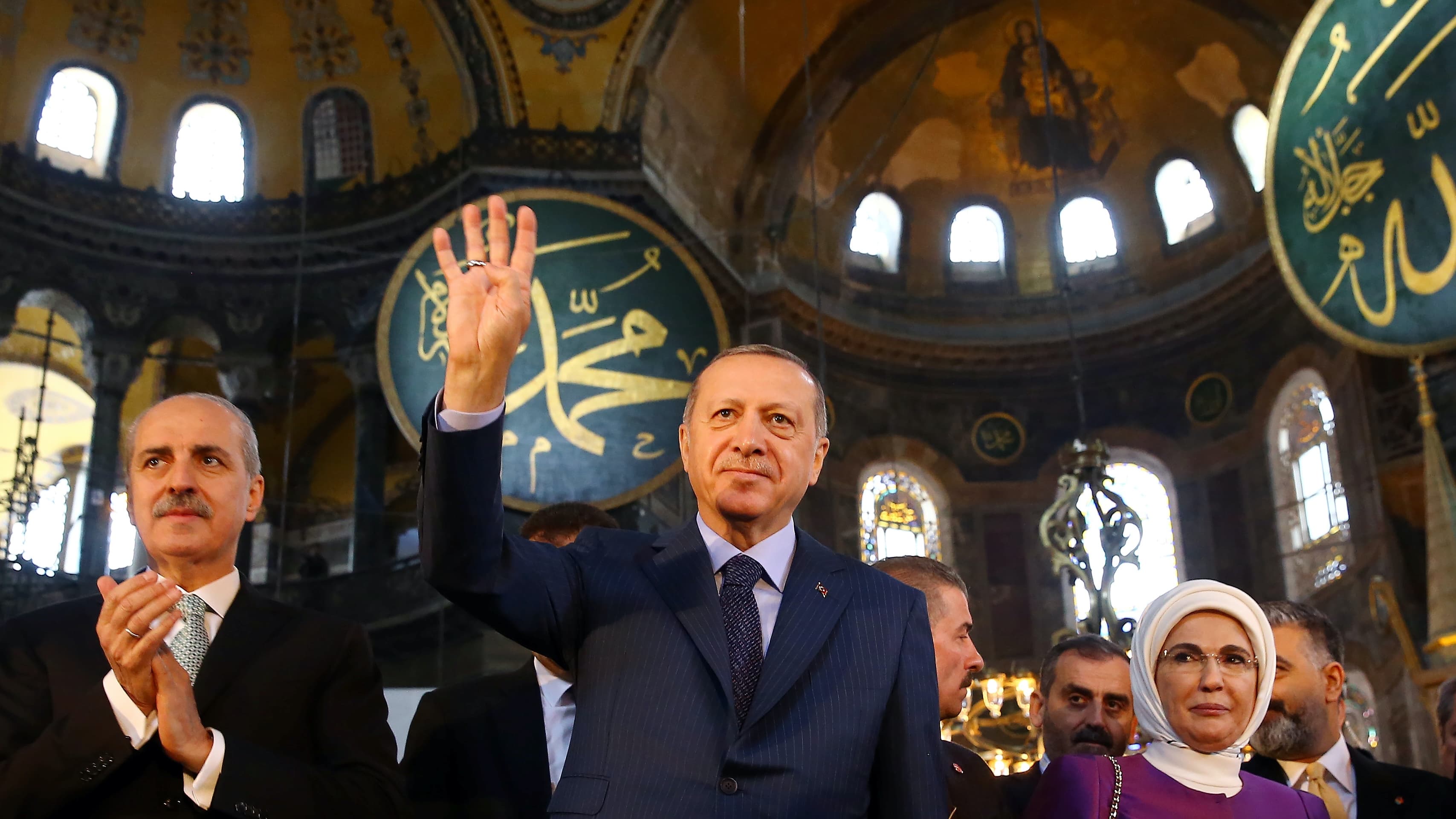A man in a suit walks with a woman wearing a dark pink dress with white head covering in ornate museum with Arabic script