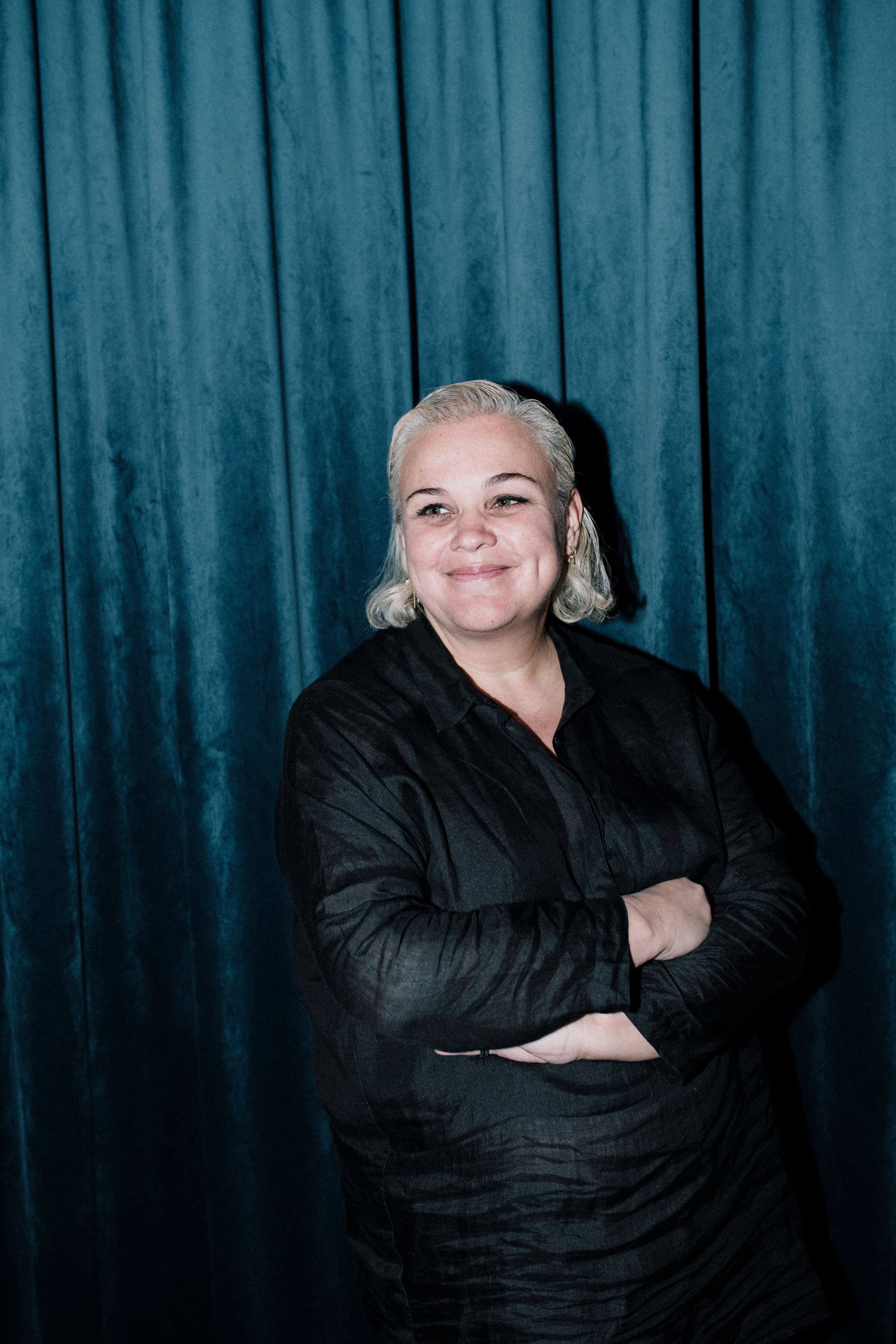 A woman with short, blonde hair wearing black dress stands in front of a blue background.