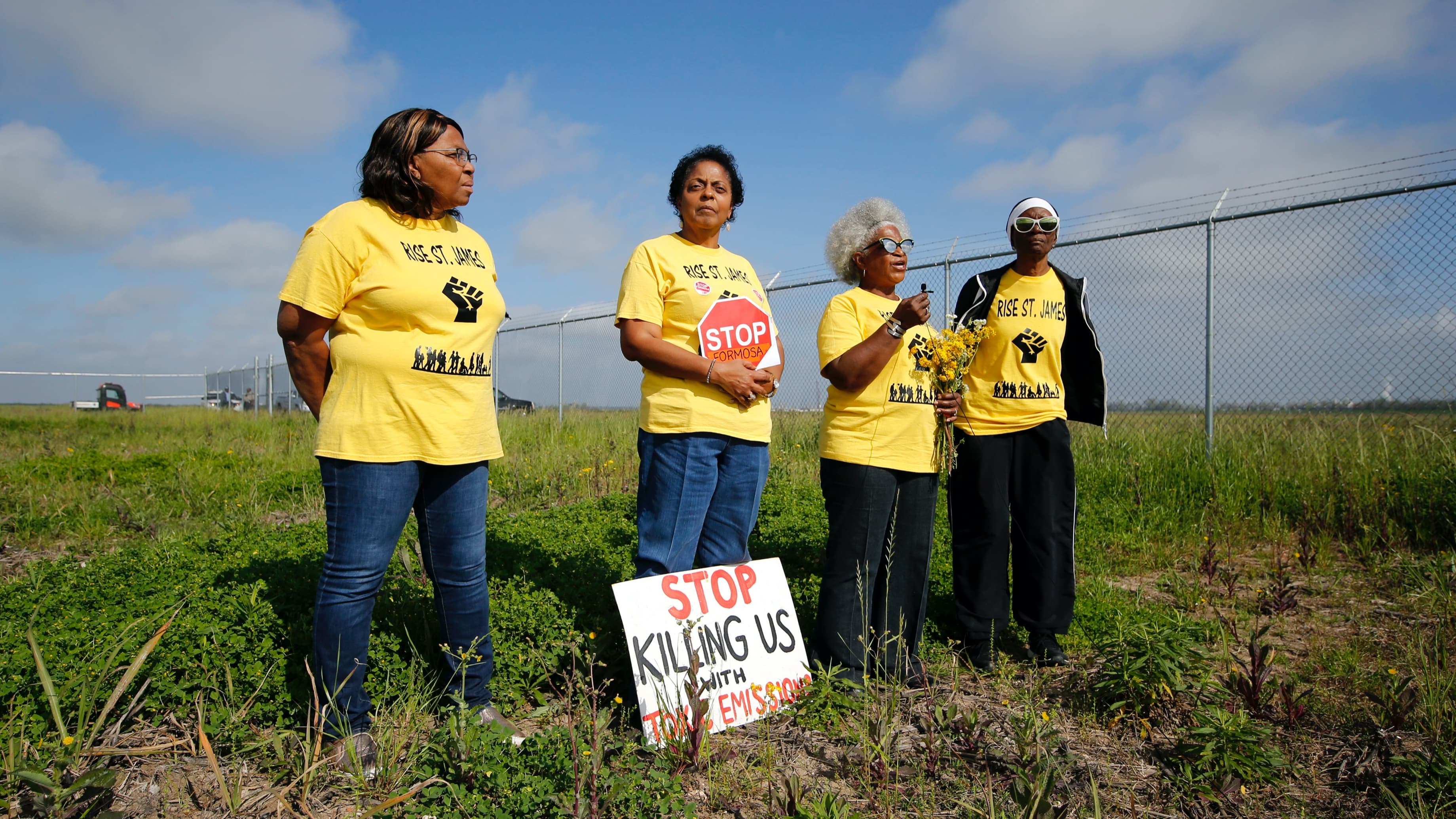 Four Black American women activists wearing yellow shirts and holding protest signs stand in a field near a fence.
