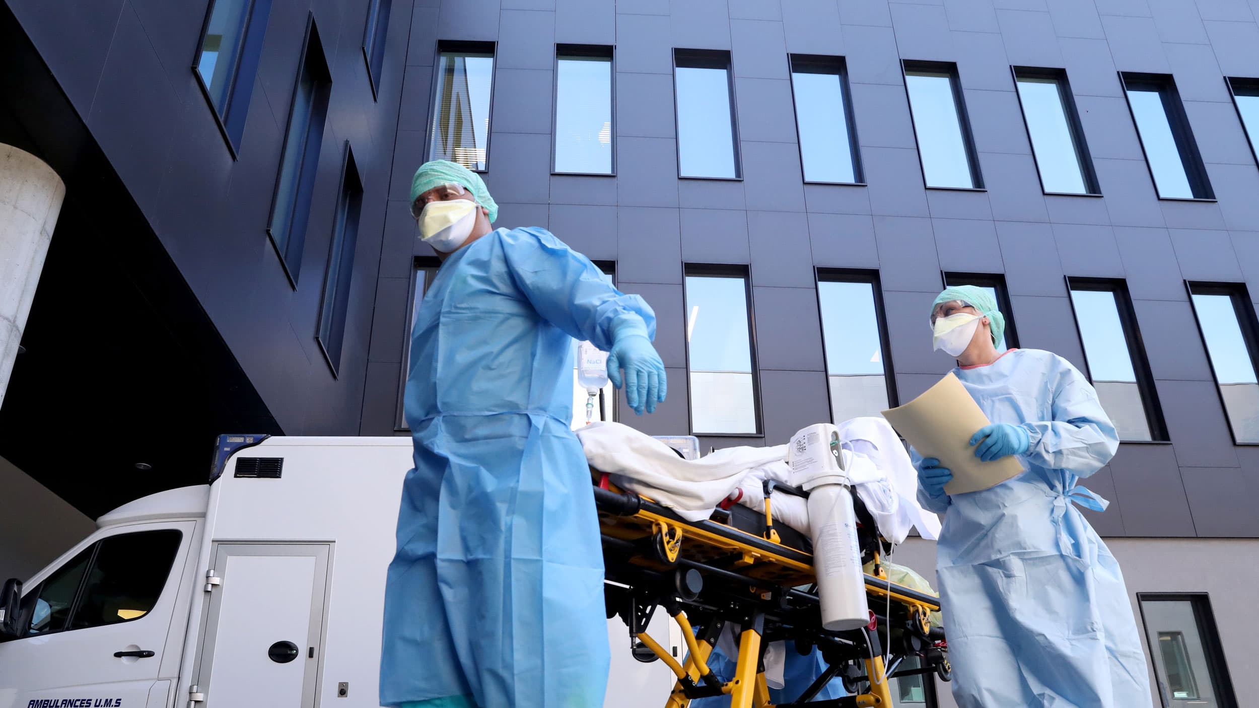 Two medical professionals are shown wearing protective gowns and face masks and pulling a gurney.