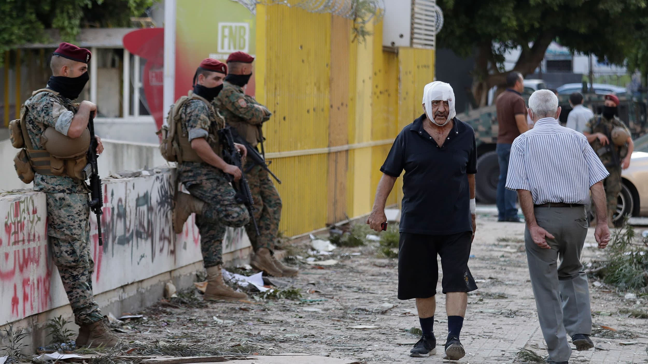 A man is shown walking on the street with a white bandage around his head with security officers standing nearby.