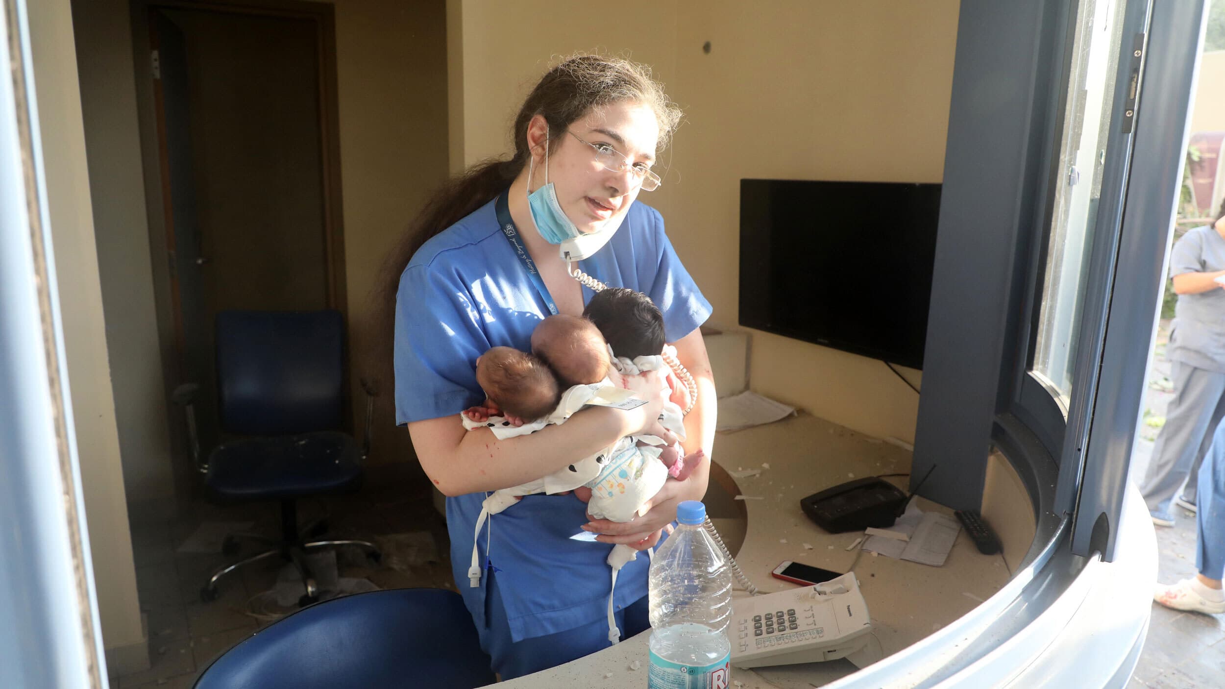 A woman with long hair and glasses wears blue scrubs holding three infants in her arms while talking on a phone in a damaged hospital