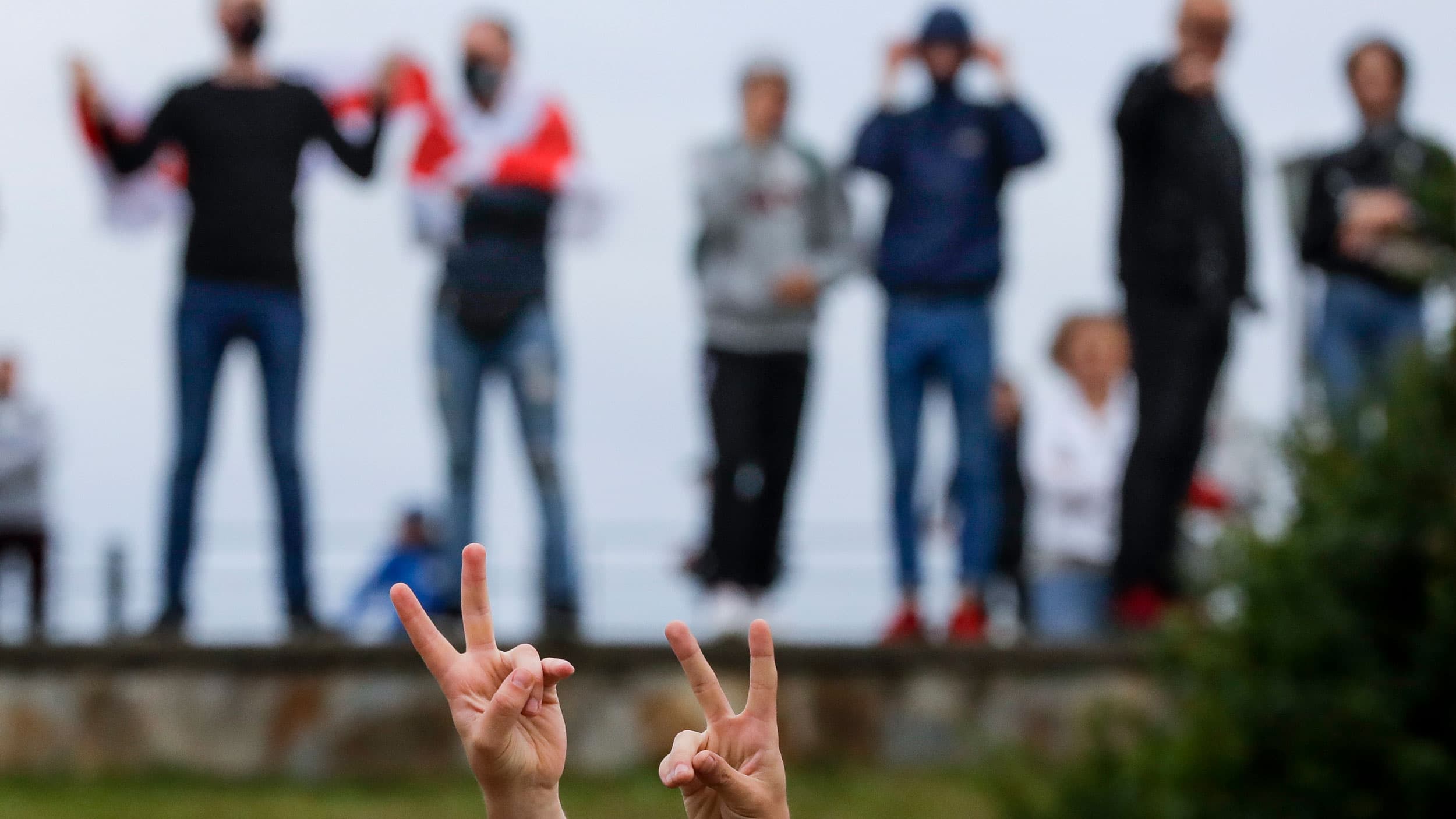 A pair of hands are shown in the nearground flashing peace signs with several people standing in a row in the background.