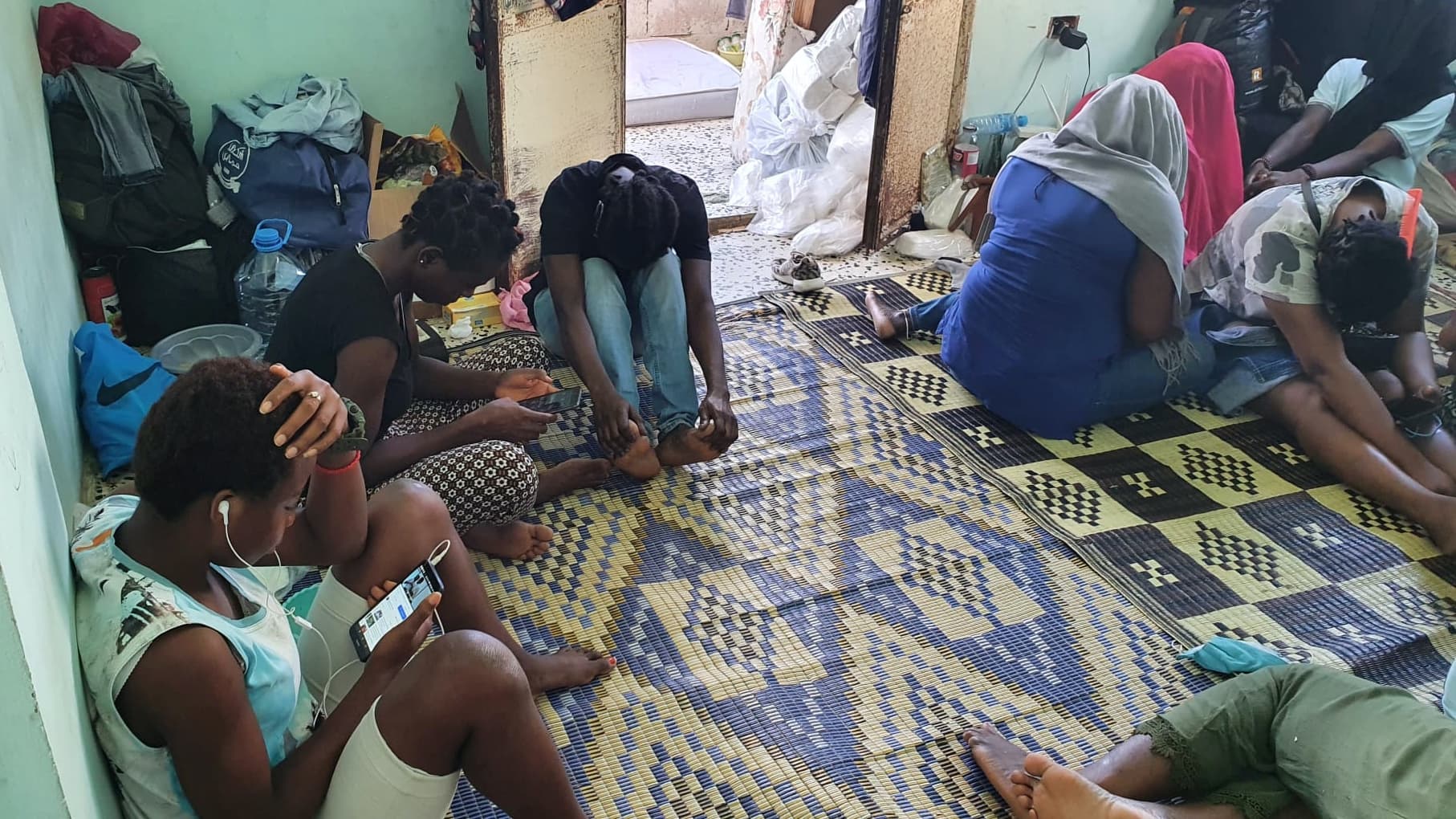 Several African women sit in a room with light blue walls, next to luggage and personal belongings.