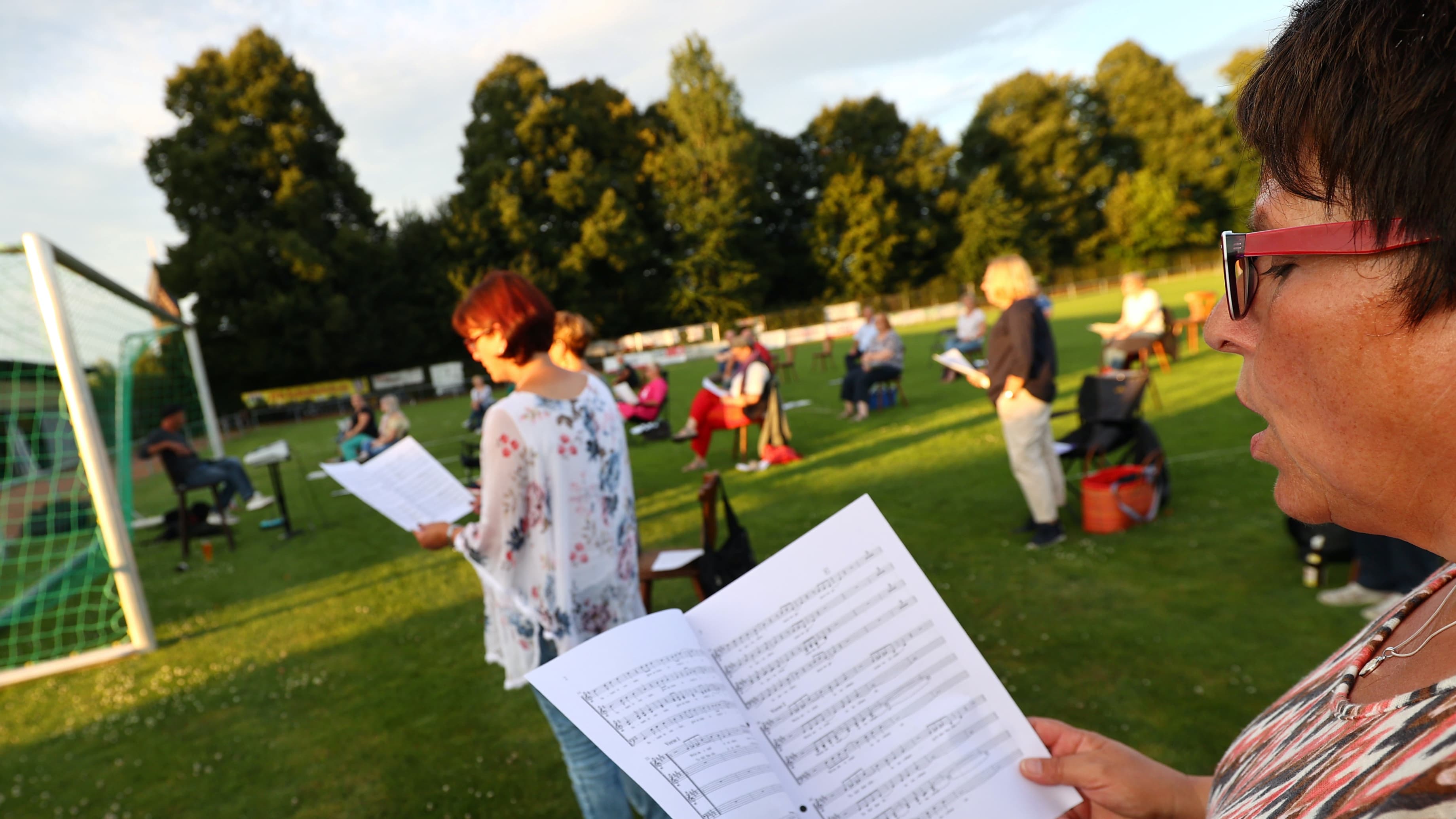 A woman sings while holding a music book in her hands in a soccer field six-feet away from another singer.