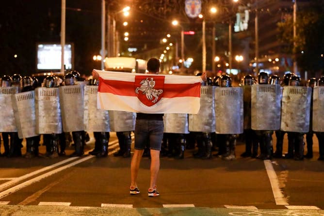 A single protestor holds up an old Belarusian national flag standing in front of a wall of officers holding large riot shields.