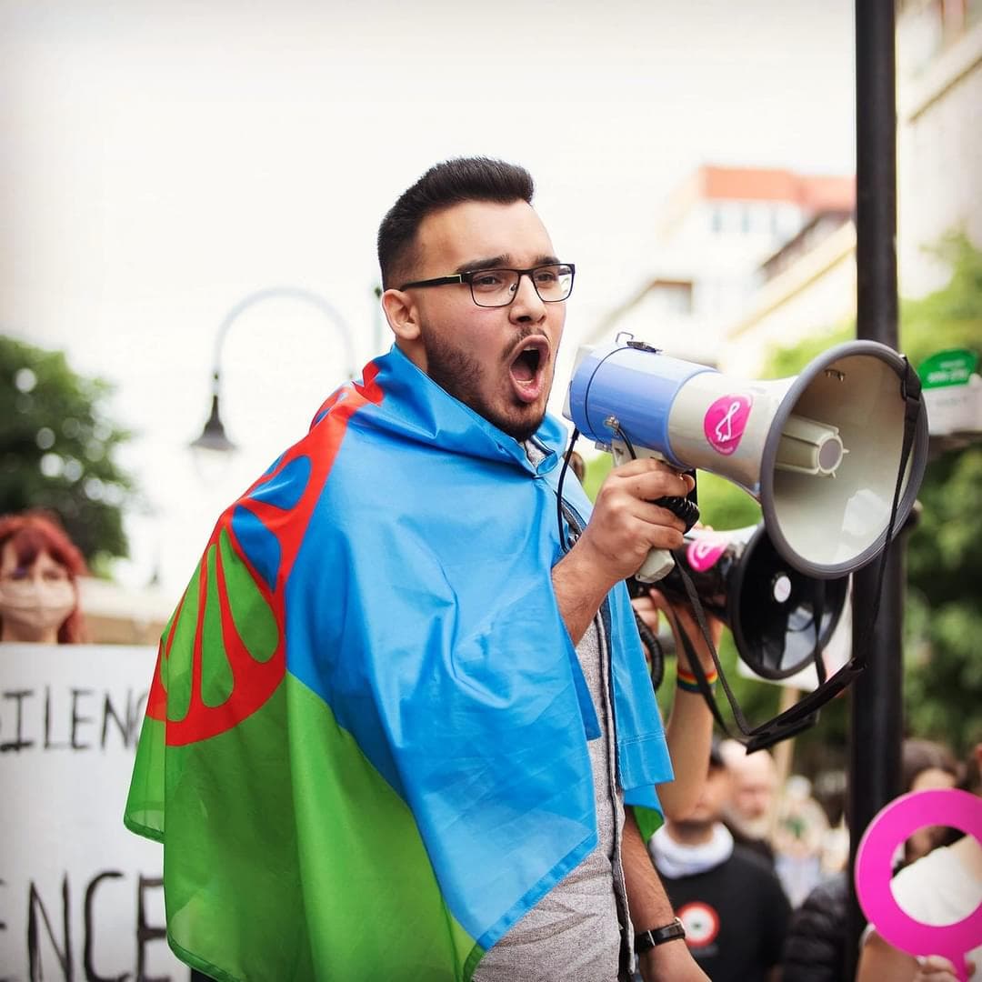 A man with glasses wears a green and blue flag draped around his shoulders.