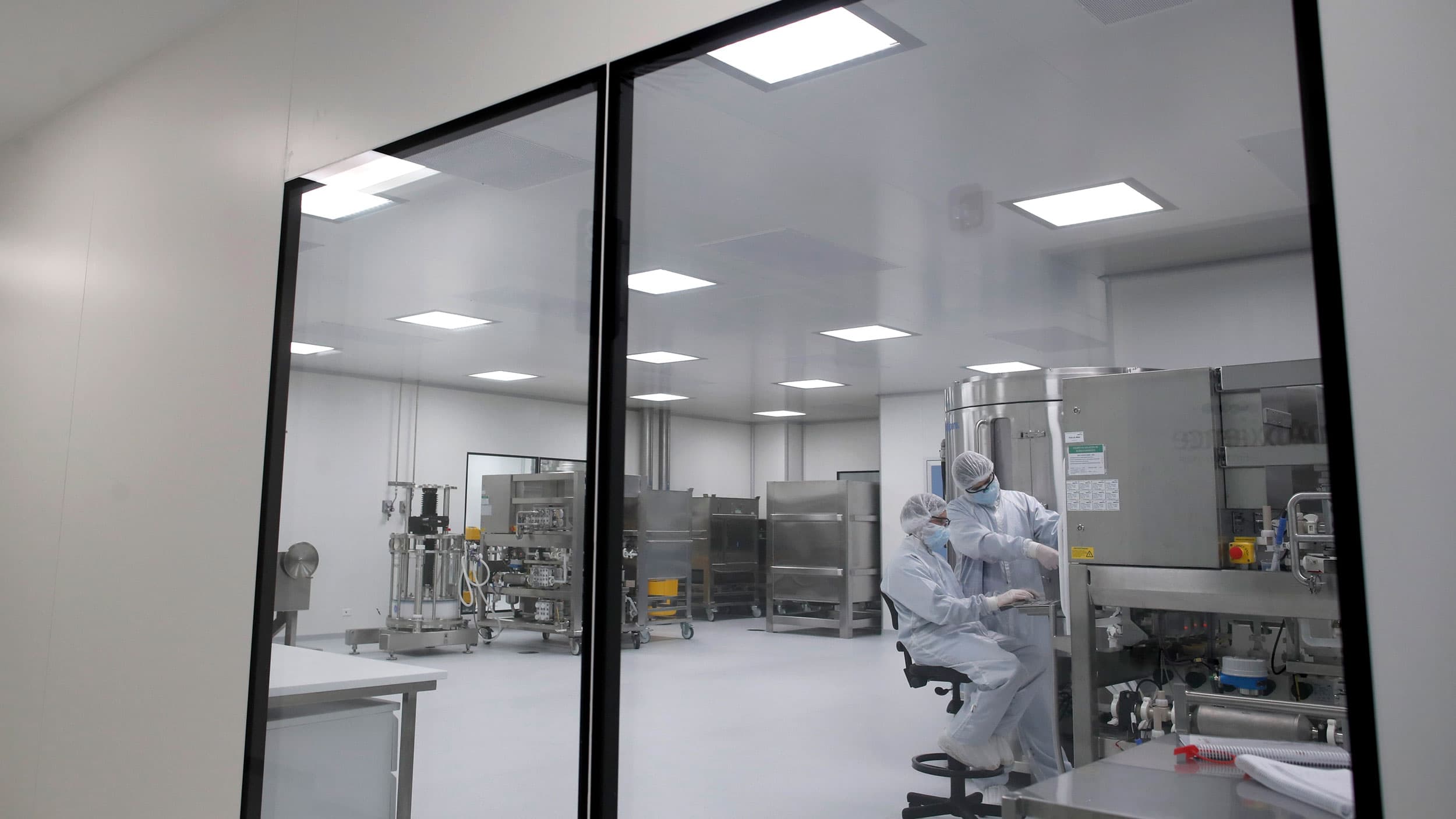 Two researchers are shown through large windows wearing full medical garments in a white laboratory.