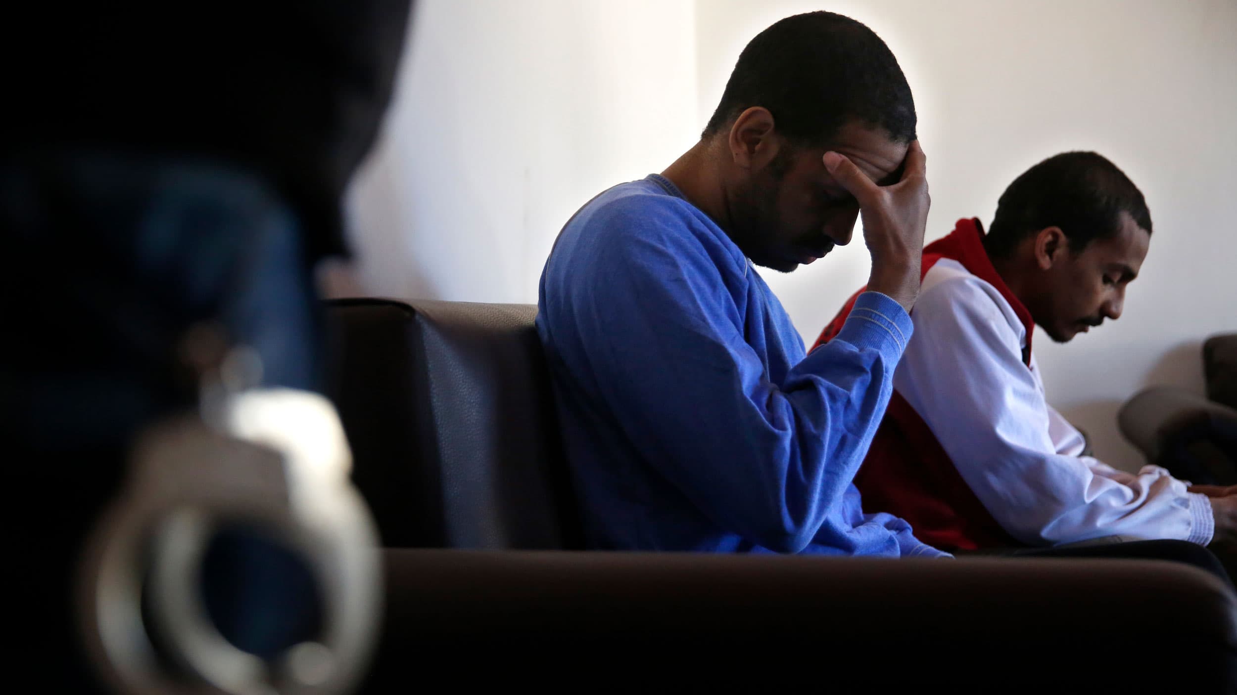 Two men are seated with one raising his hand to his face and a pair of handcuffs shown in soft focus in the nearground.