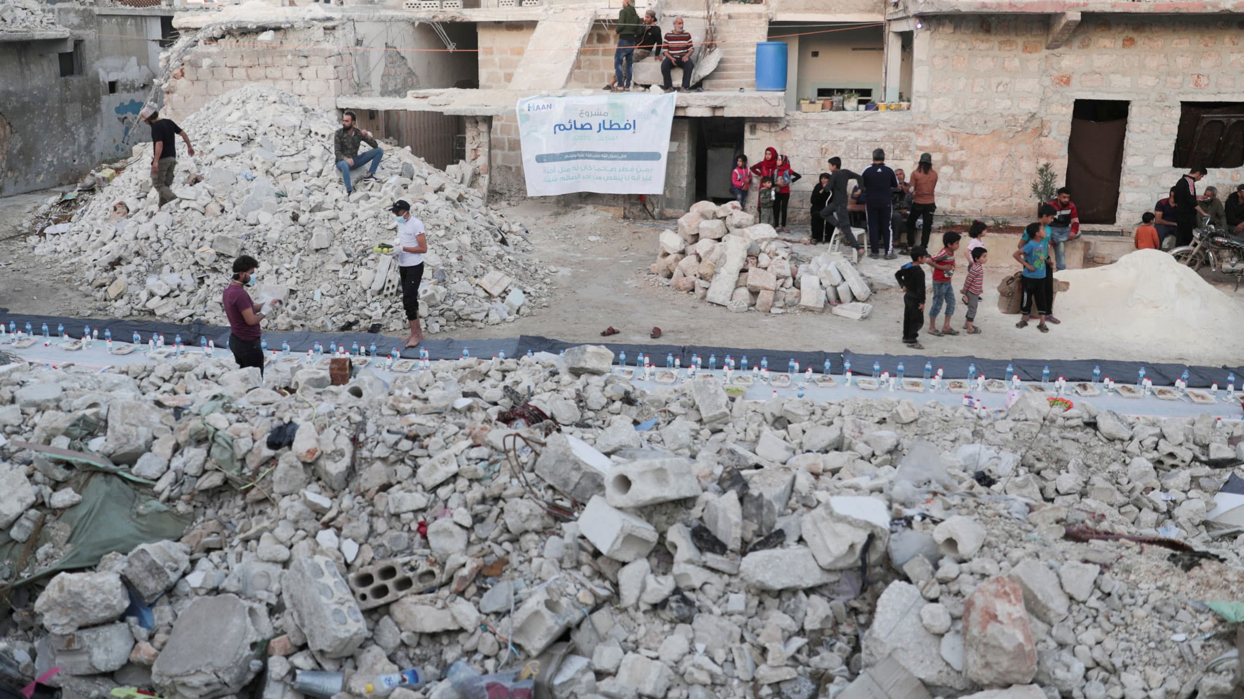 Several people are shown preparing meals next to a large pile of rubble and a line of water bottles.