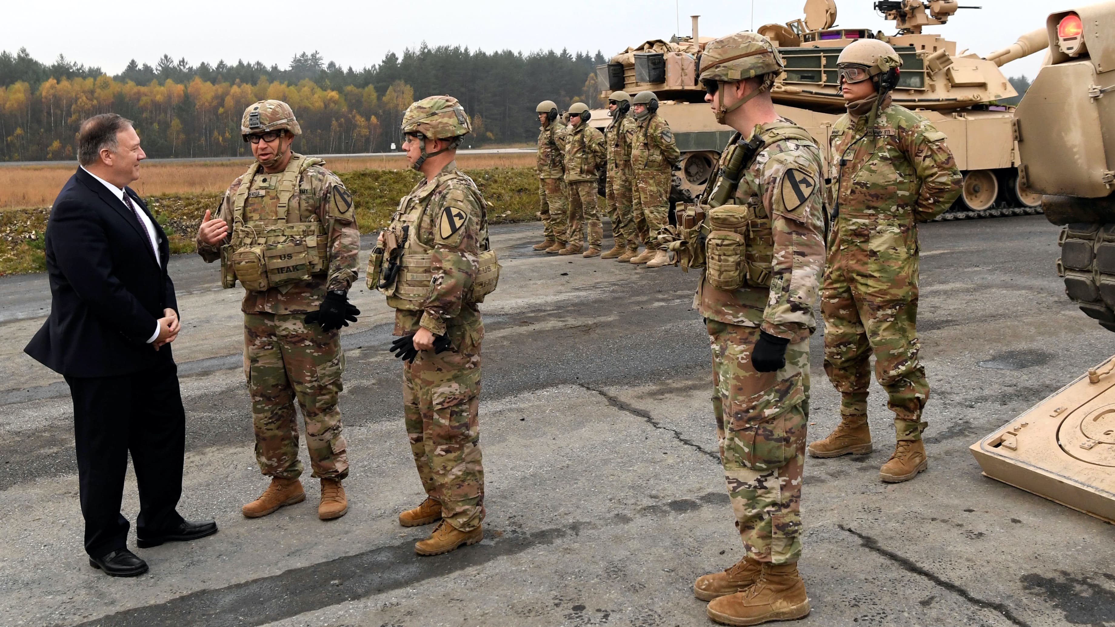 A man in a dark suit talks with men wearing military uniforms outside near tan tanks.