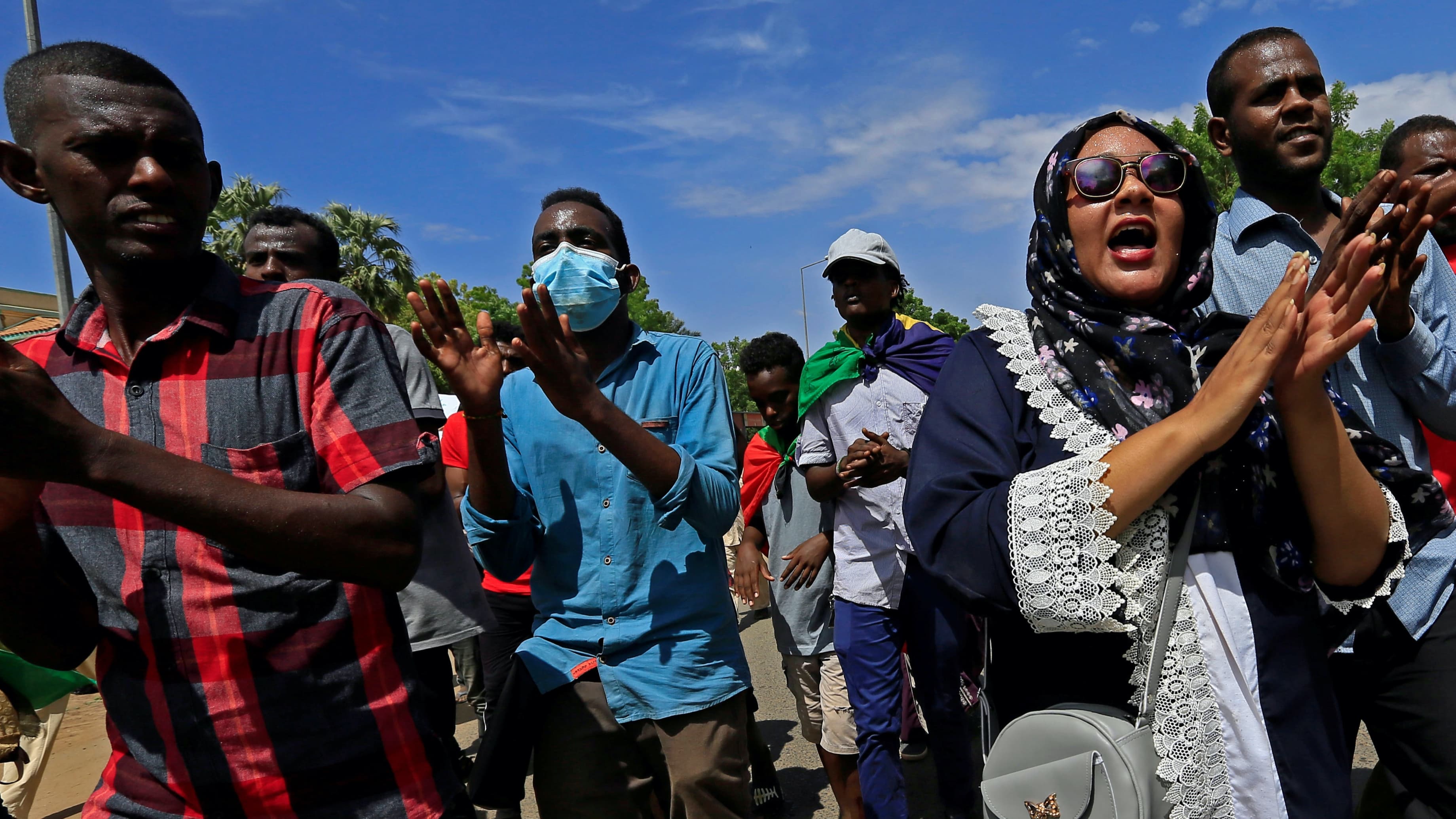 Protesters clap and cheer on the streets under a hot sun and blue sky, with one woman wearing sunglasses and head covering