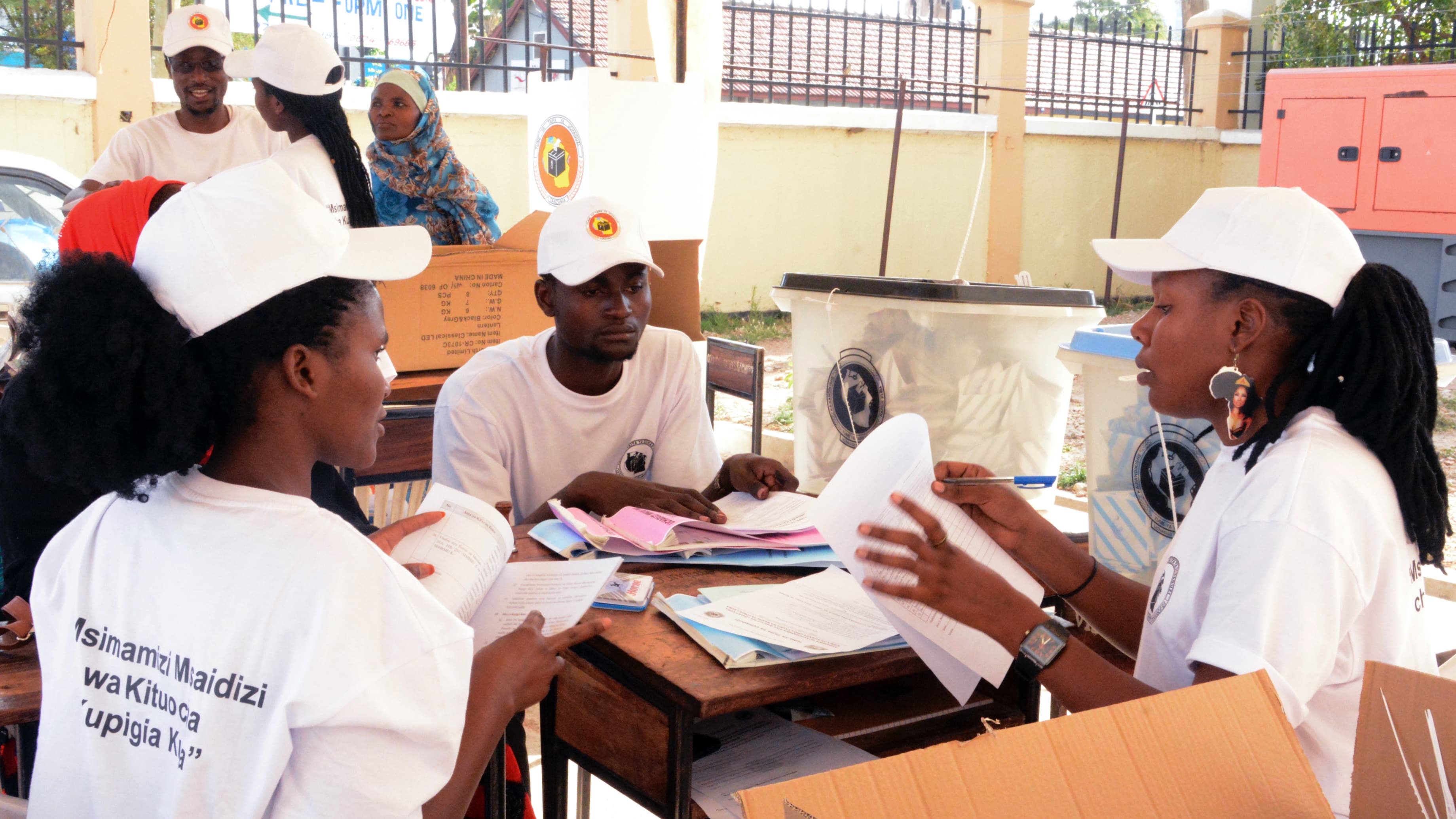 Three poll workers wearing white shirts and hats sit at a table together and sort through papers