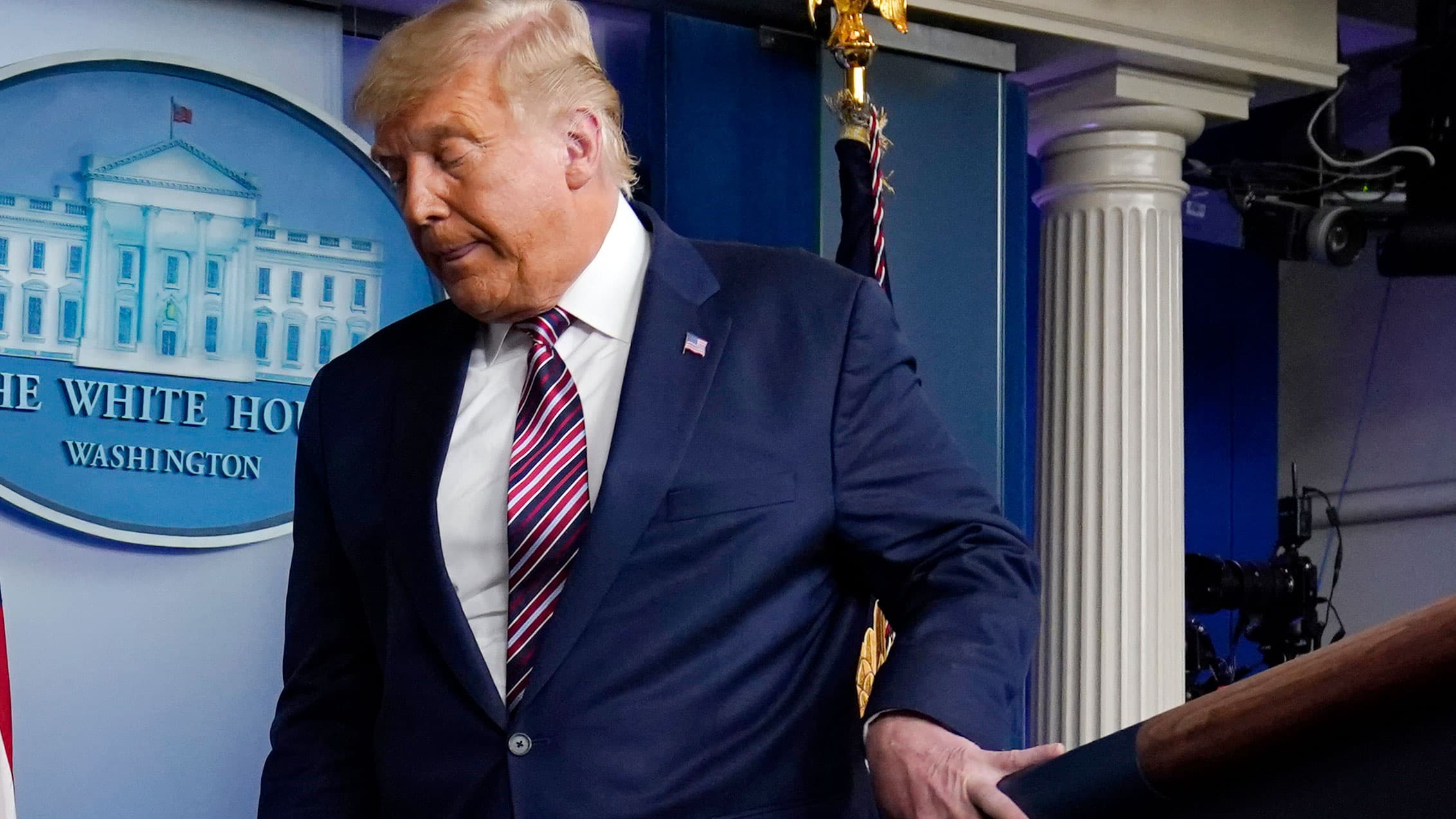 President Donald Trump is shown wearing a blue suit and stripped tie while turning and walking away from a podium.