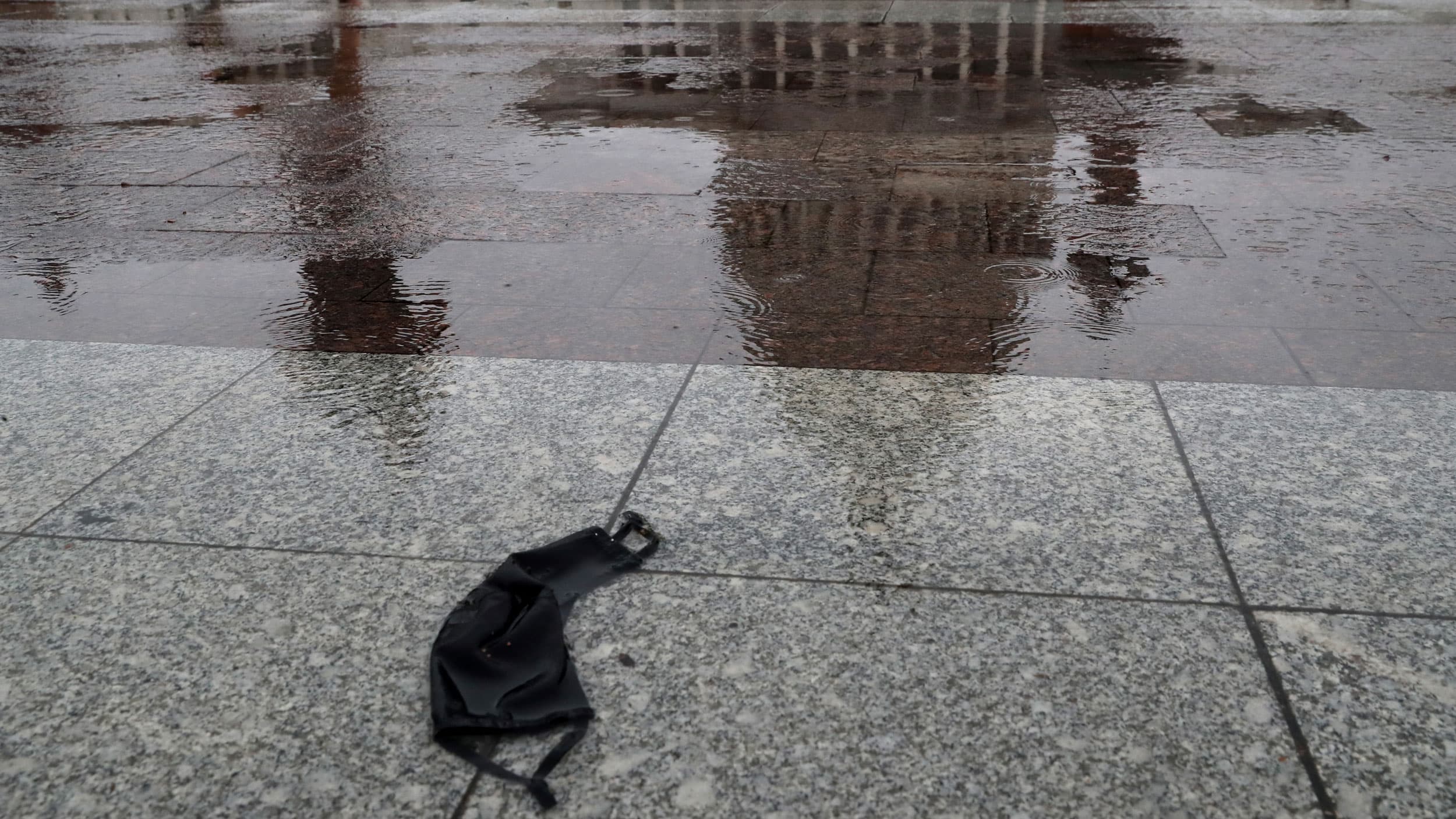 An abandoned mask, strewn on the wet stone ground next to a puddle with the reflection of the US capitol building.